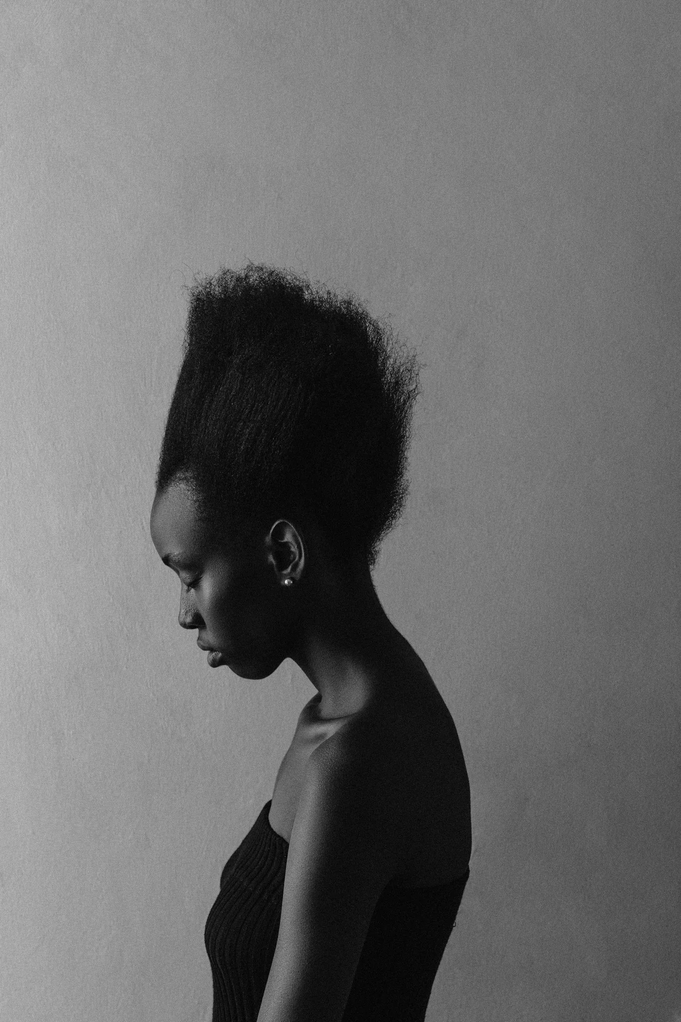 A high-contrast, black-and-white fine art side profile of a black female model with a tall natural afro hairstyle, wearing a sleeveless black top, against a plain light-coloured wall.
