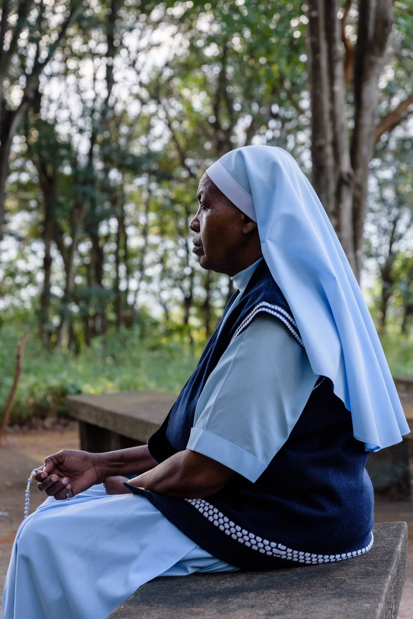 A nun sitting outdoors on a stone surface, holding a rosary, praying, with trees in the background at the Usambara Sister Convent in Korogwe, Tanzania.