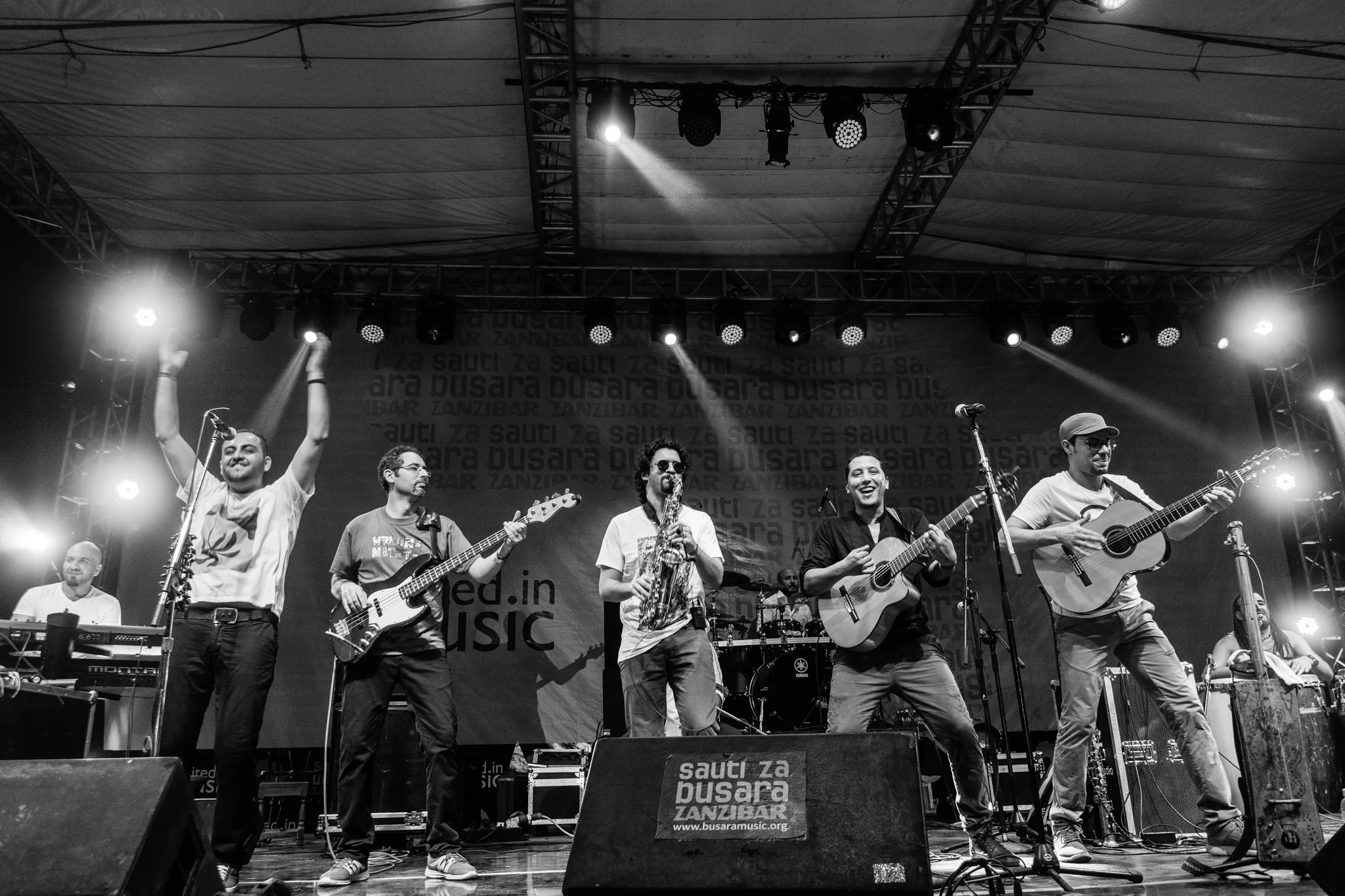 A live music performance features a band on stage with six members playing various instruments, including guitars, bass, keyboard, saxophone, drums, and percussion, under stage lighting in Zanzibar at the Sauti za Busara Music Festival in 2018.