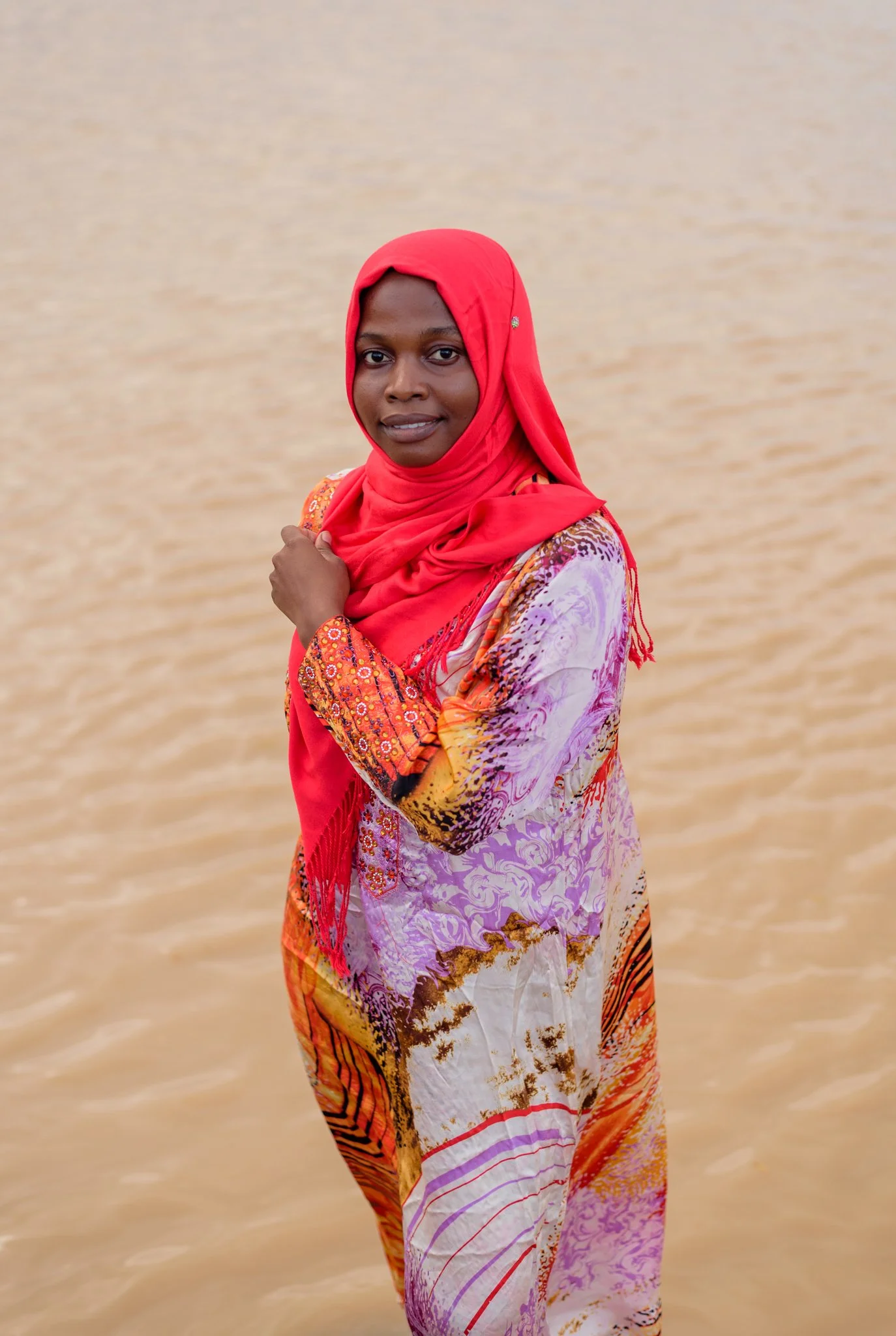 A woman wearing a vibrant hijab and colorful dress standing in shallow water.
