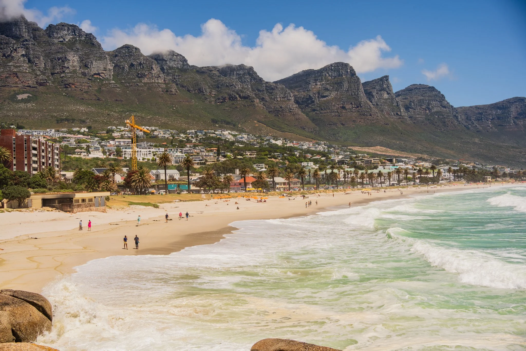 View of a sandy beach with waves coming ashore, bordered by palm trees. Behind the beach are buildings and a mountain range with clouds.