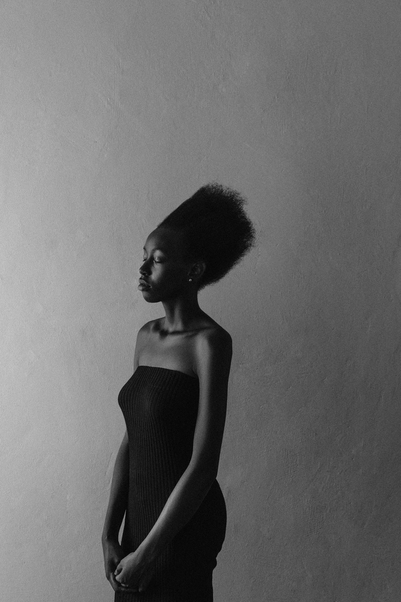 A high-contrast, black-and-white fine art photo of a black female model with natural hair wearing a strapless dress, standing with her eyes closed against a plain wall.