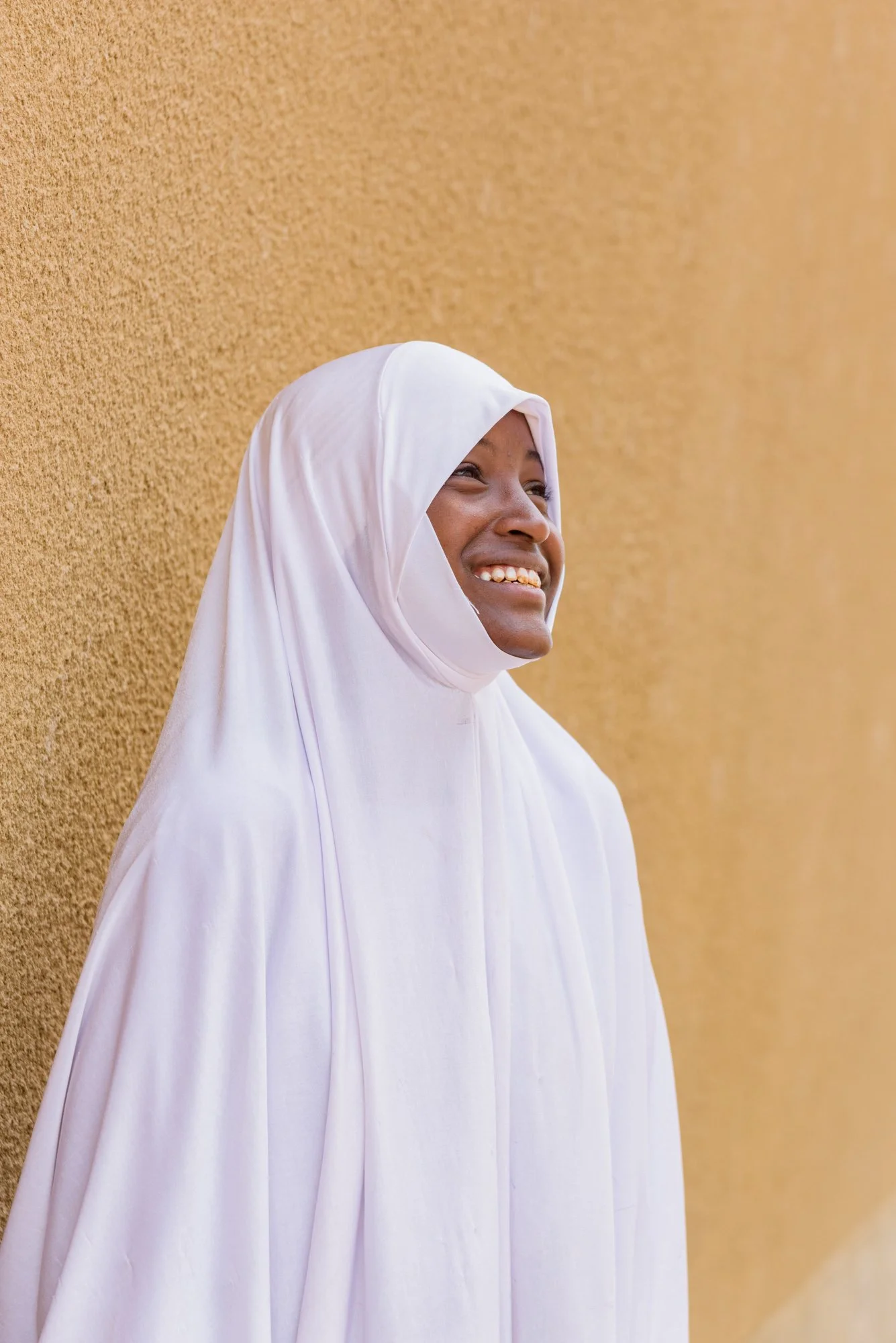 Girl wearing a white hijab and dress smiles while standing against a textured yellow wall in Arusha, Tanzania.