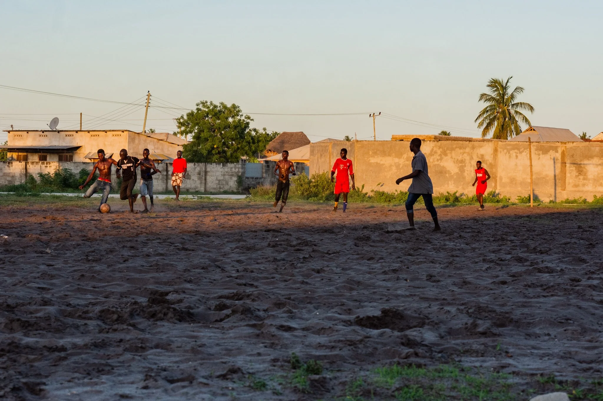 Men playing football on a sandy field in a neighbourhood with houses and trees in the background during late afternoon in Kigamboni, Dar es Salaam.