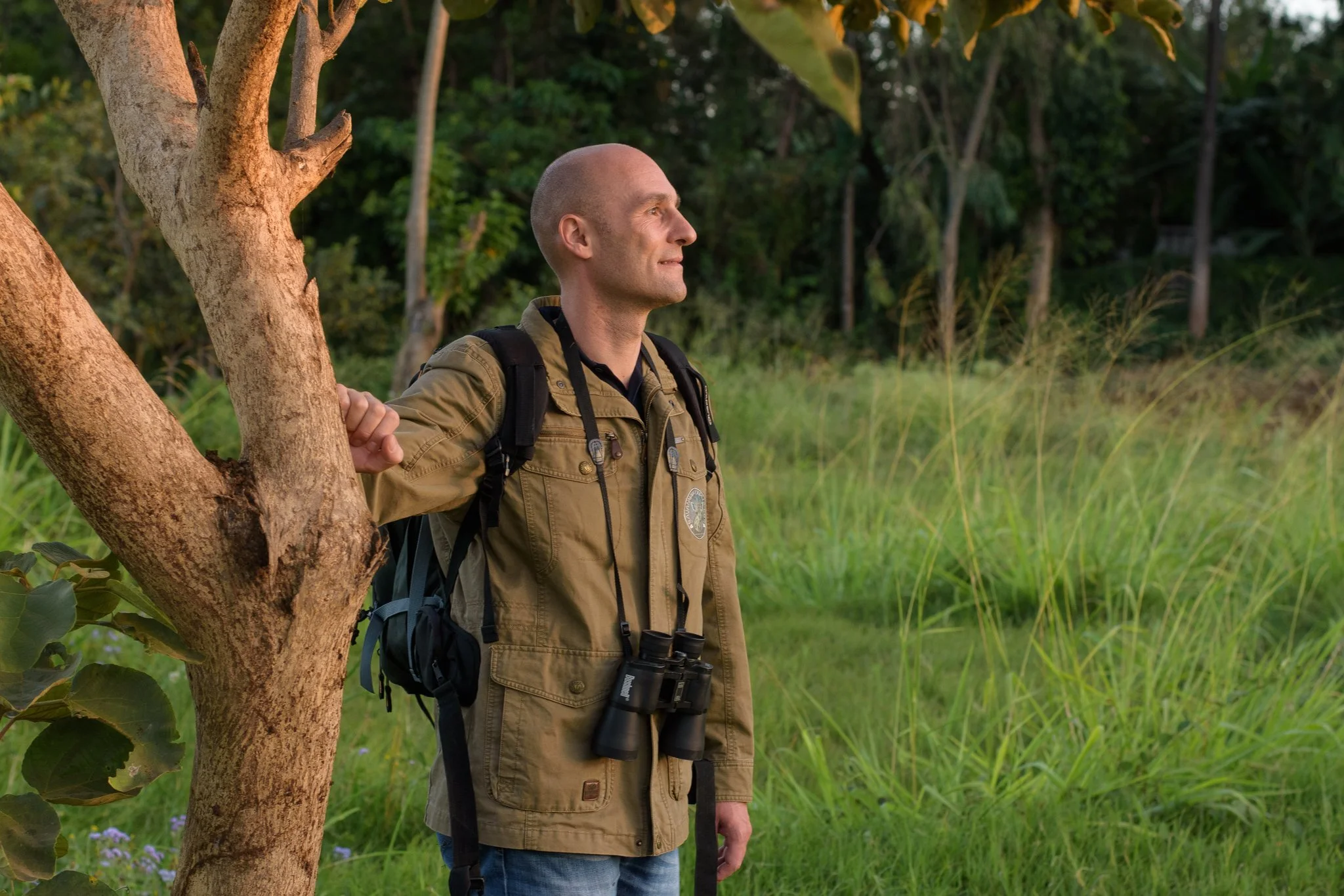 A man wearing a tan jacket and backpack, with binoculars hanging around his neck, standing next to a tree in a grassy field, looking to the right.