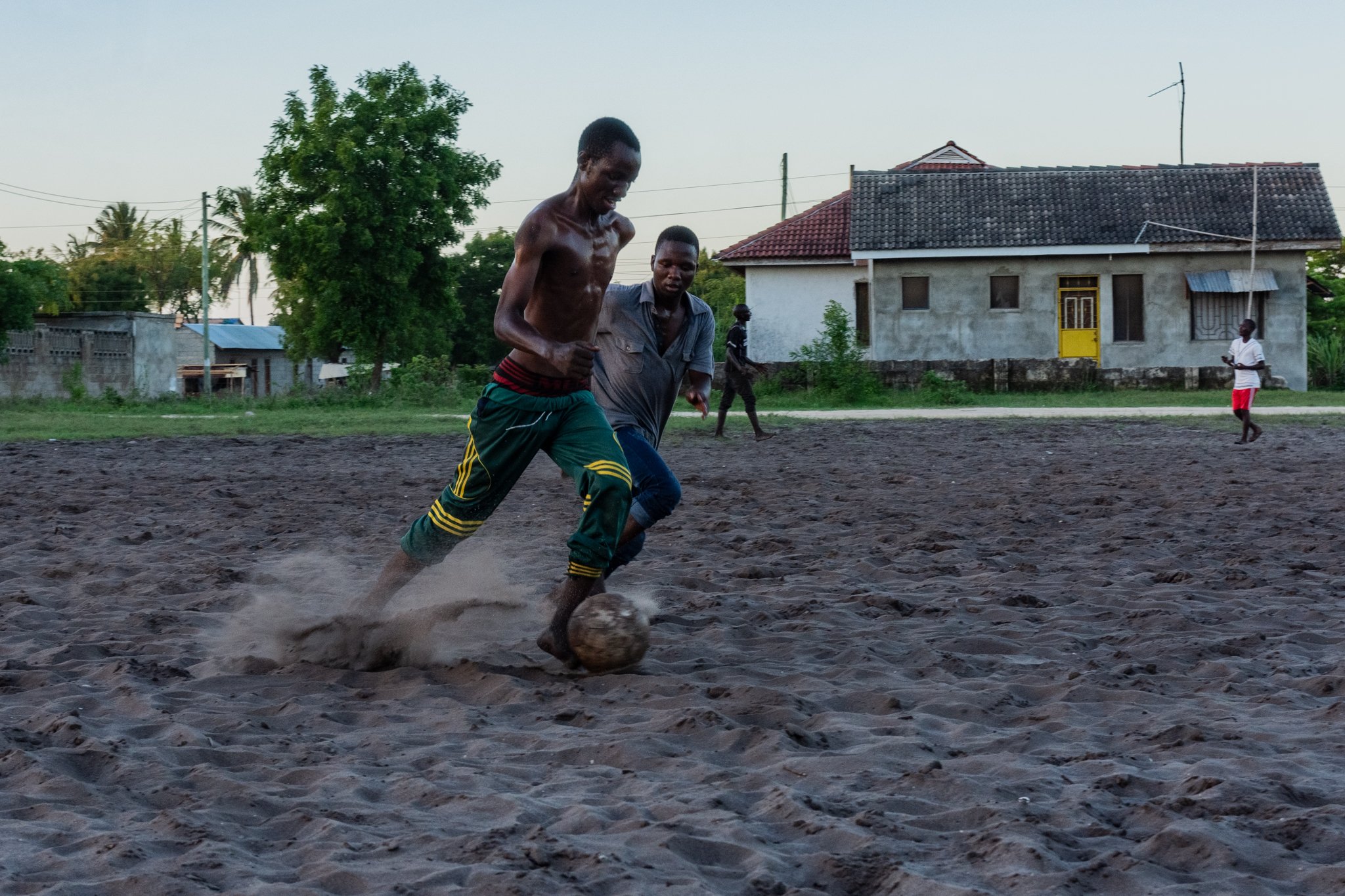 Two men playing football on a sandy field, with three other people and houses in the background in Kigamboni, Dar es Salaam.