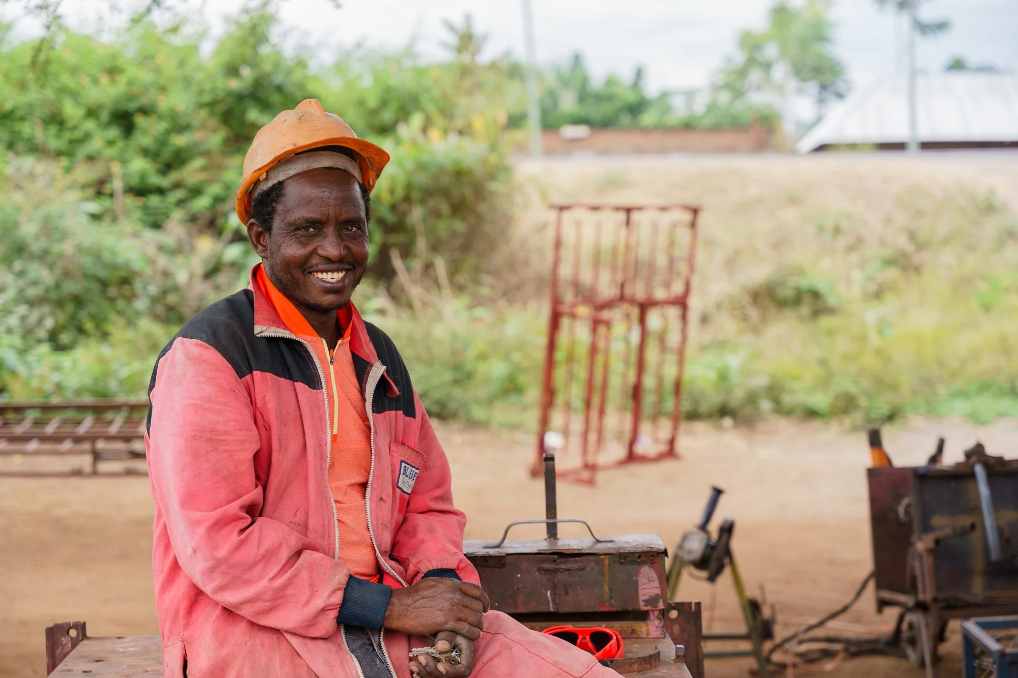 A smiling man wearing a construction helmet and work clothes sits outdoors with tools and equipment around him in Babati, Manyara, Tanzania.