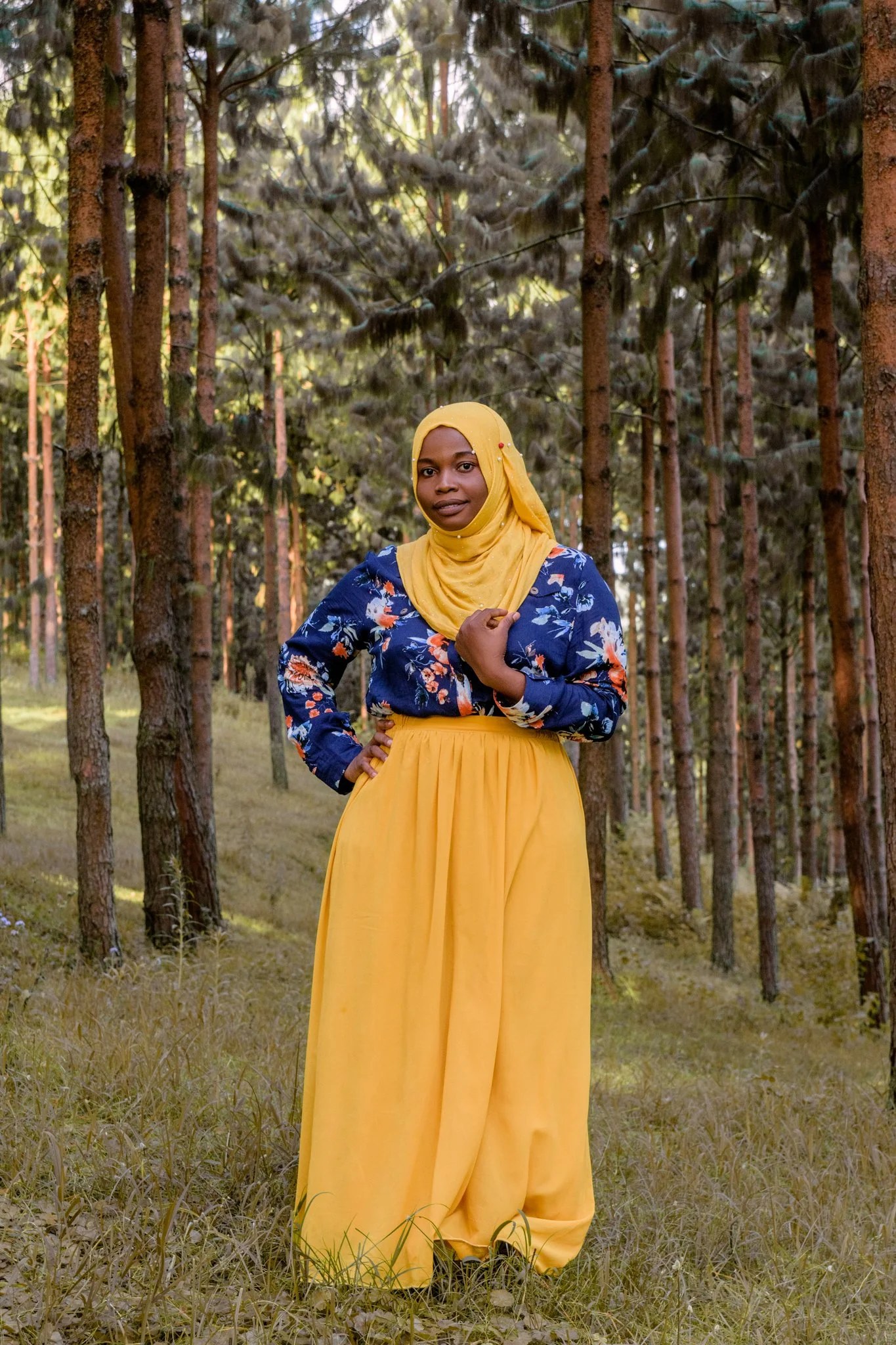 A woman wearing a yellow hijab and a long yellow skirt with a blue floral blouse stands in a forest with tall trees and green grass.