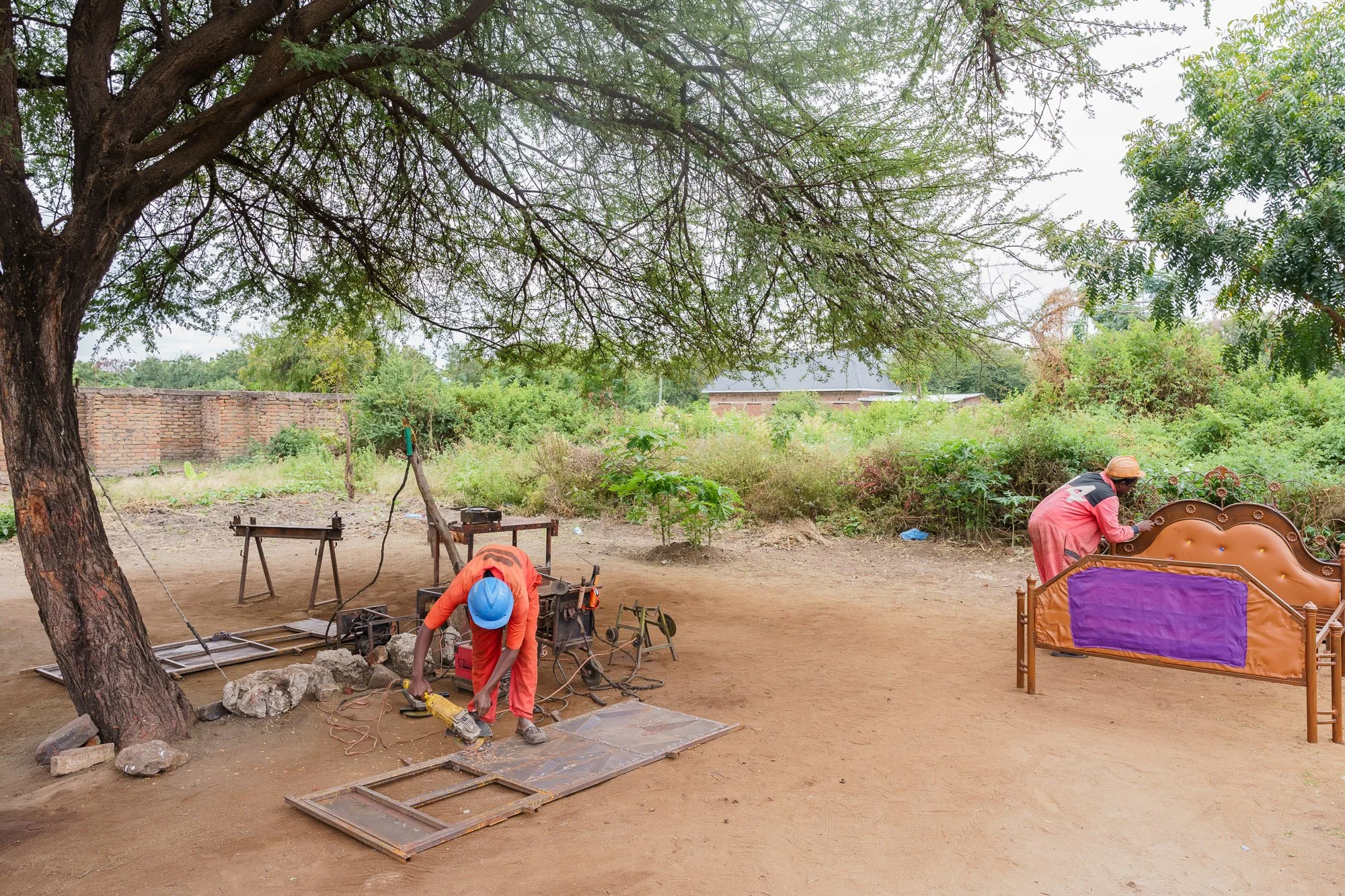 Two workers in safety helmets assembling furniture outdoors under a large tree in Babati, Manyara, Tanzania.
