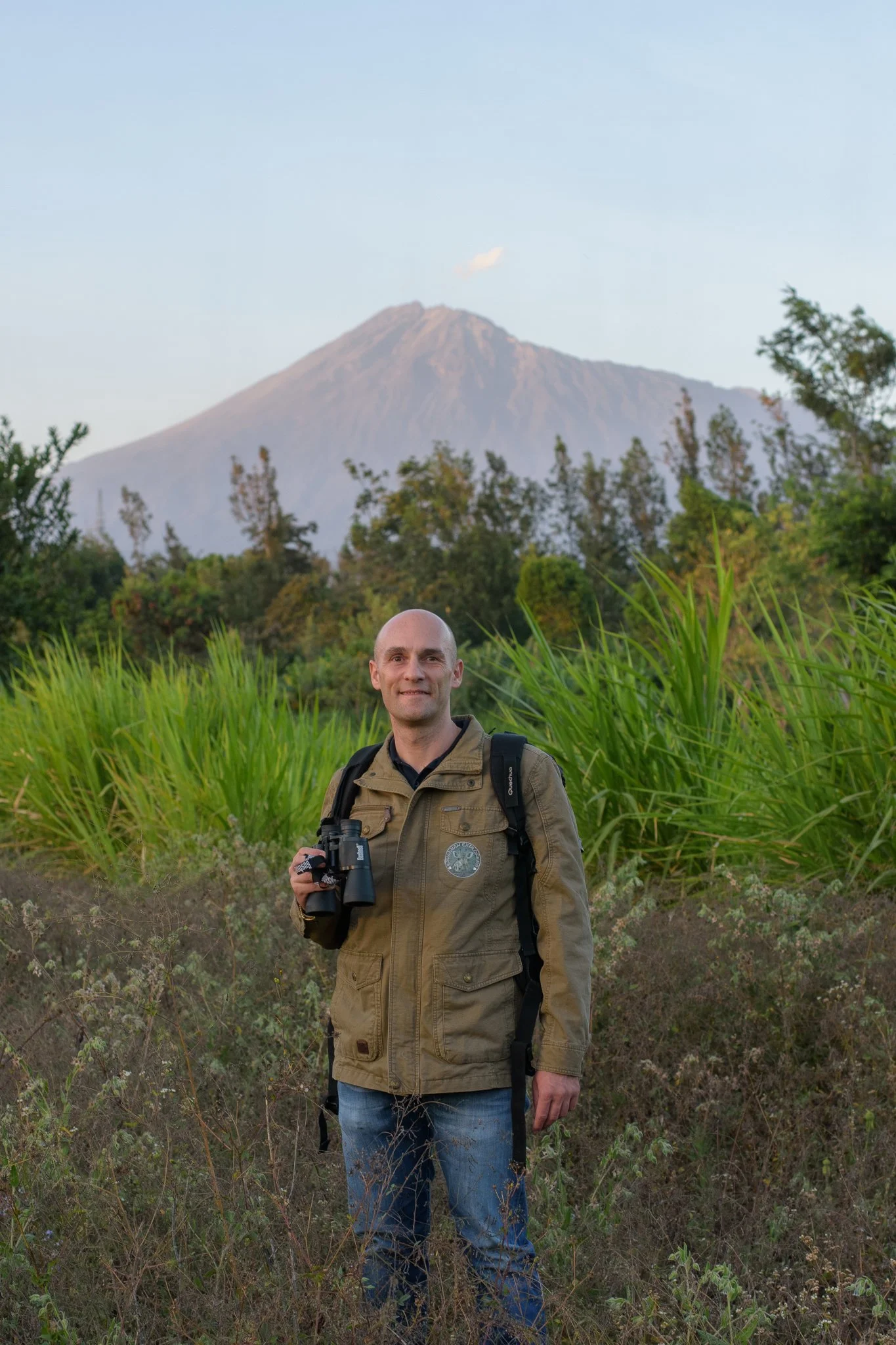 A man standing outdoors in front of a lush green landscape with a mountain in the background, holding binoculars in Arusha, Tanzania.