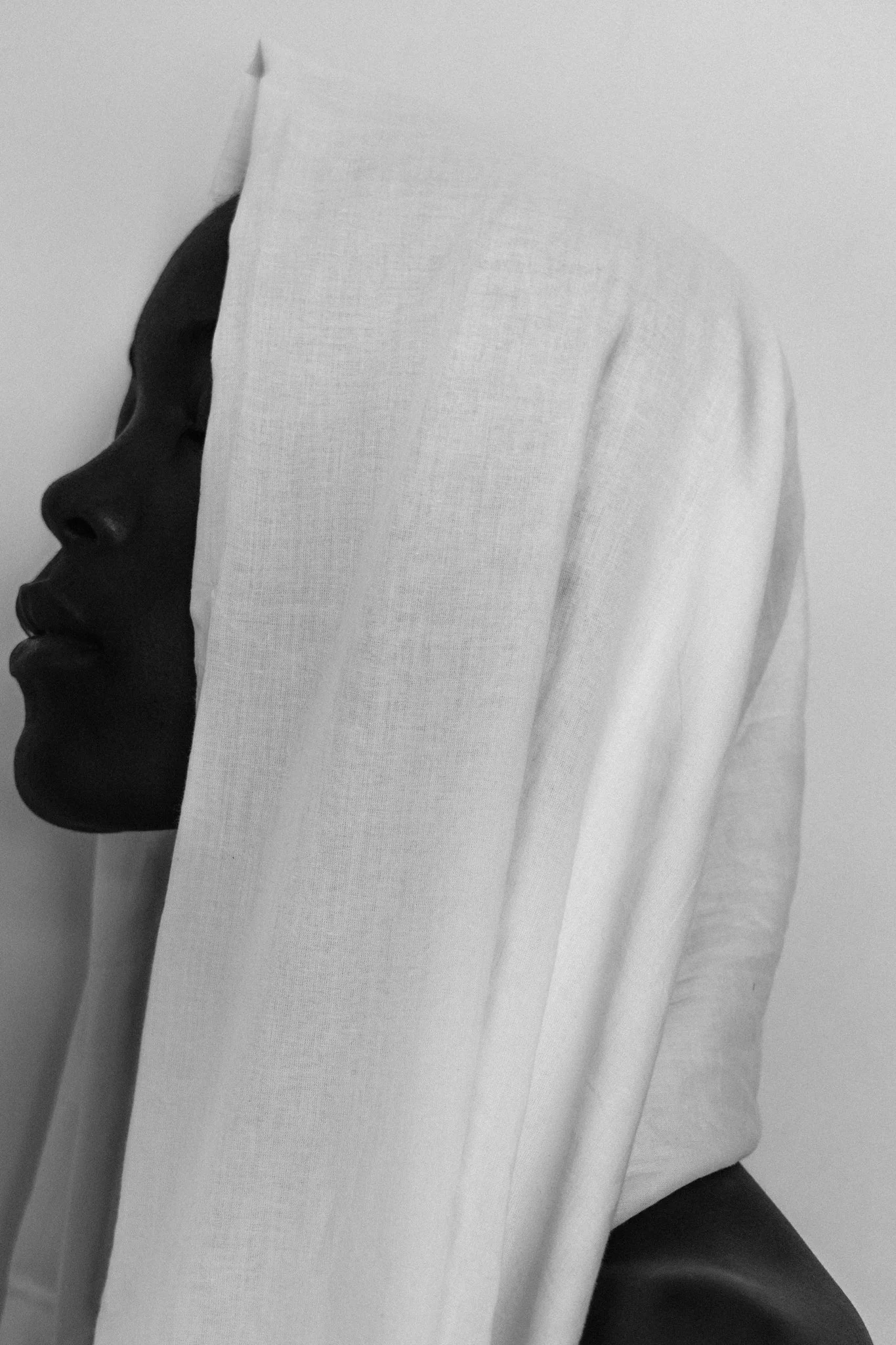 A high-contrast, black-and-white fine art side-profile portrait of a female model with a dark skin tone, covered in white fabric, against a plain background in Dar es Salaam, Tanzania.