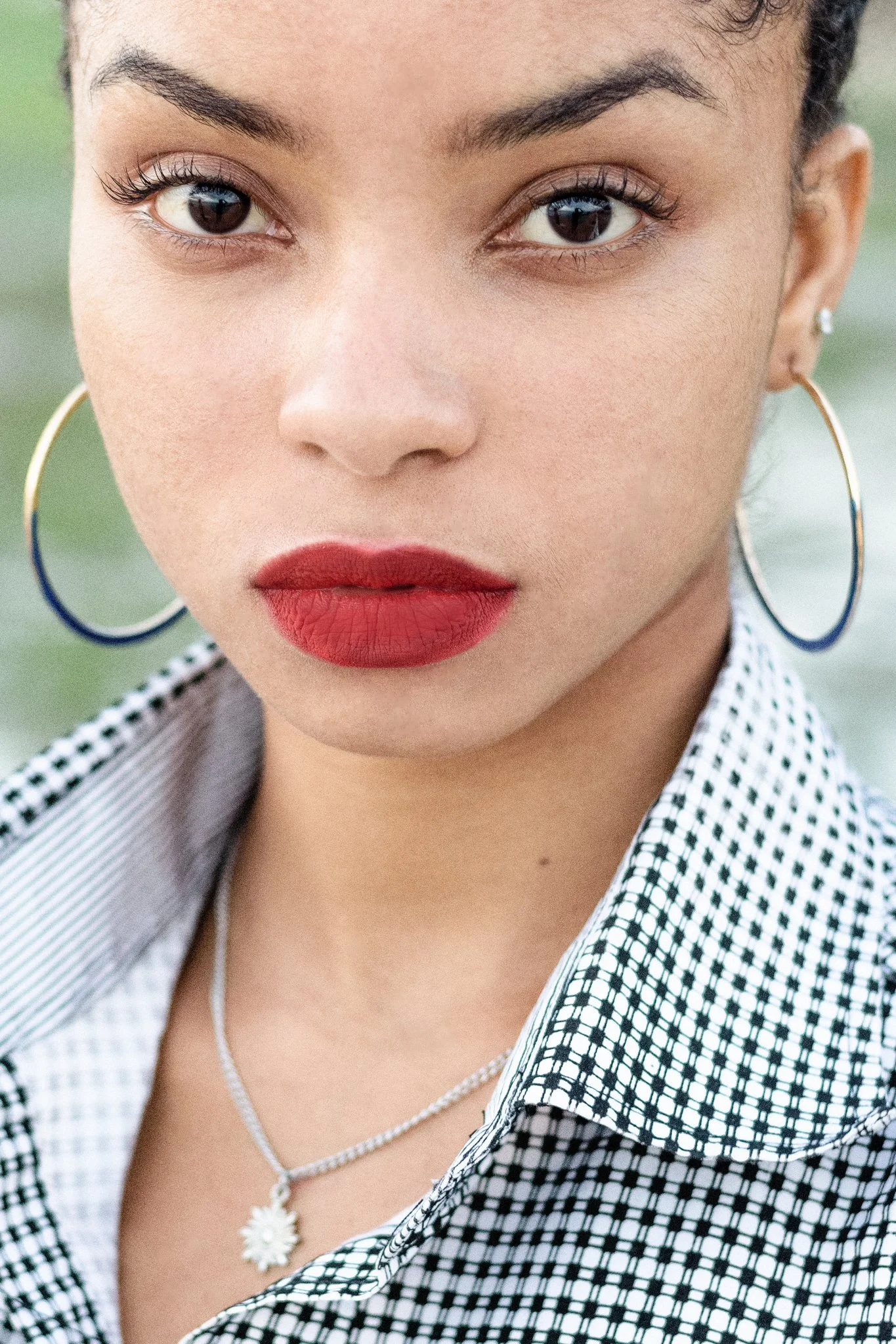 Close-up portrait of a woman with dark hair, large hoop earrings, red lipstick, and a black-and-white checkered shirt, outdoors with blurred green background.