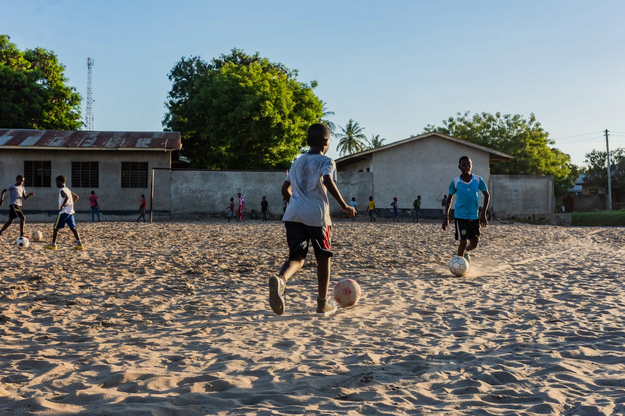 Children playing football on a sandy field with houses and trees in the background in Mbagala, Dar es Salaam.