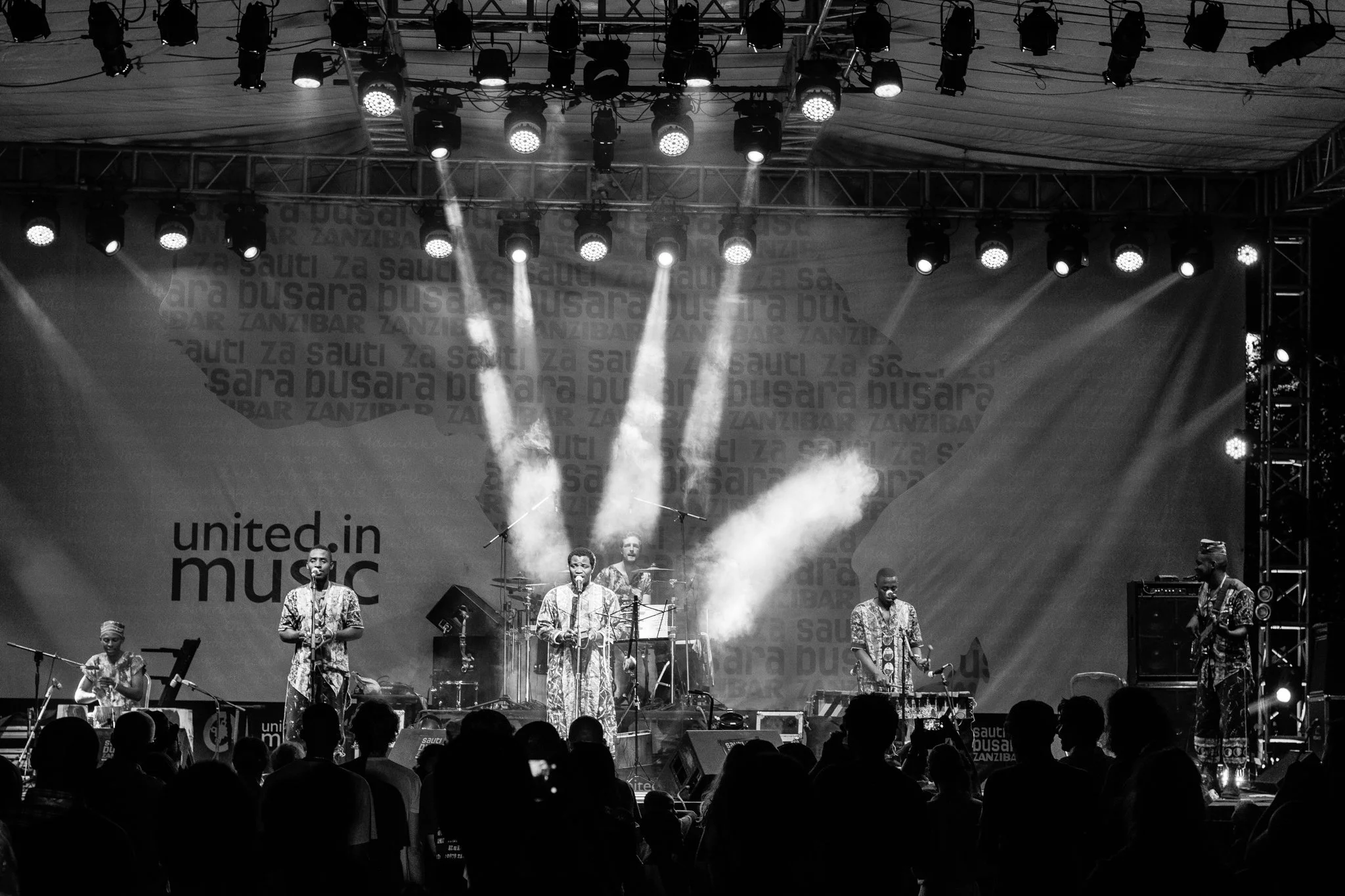 A live musical performance on stage with several musicians, some playing instruments, under stage lighting and fog effects. Audience members are visible in the foreground at the Sauti za Busara Music Festival in Zanzibar in 2018.