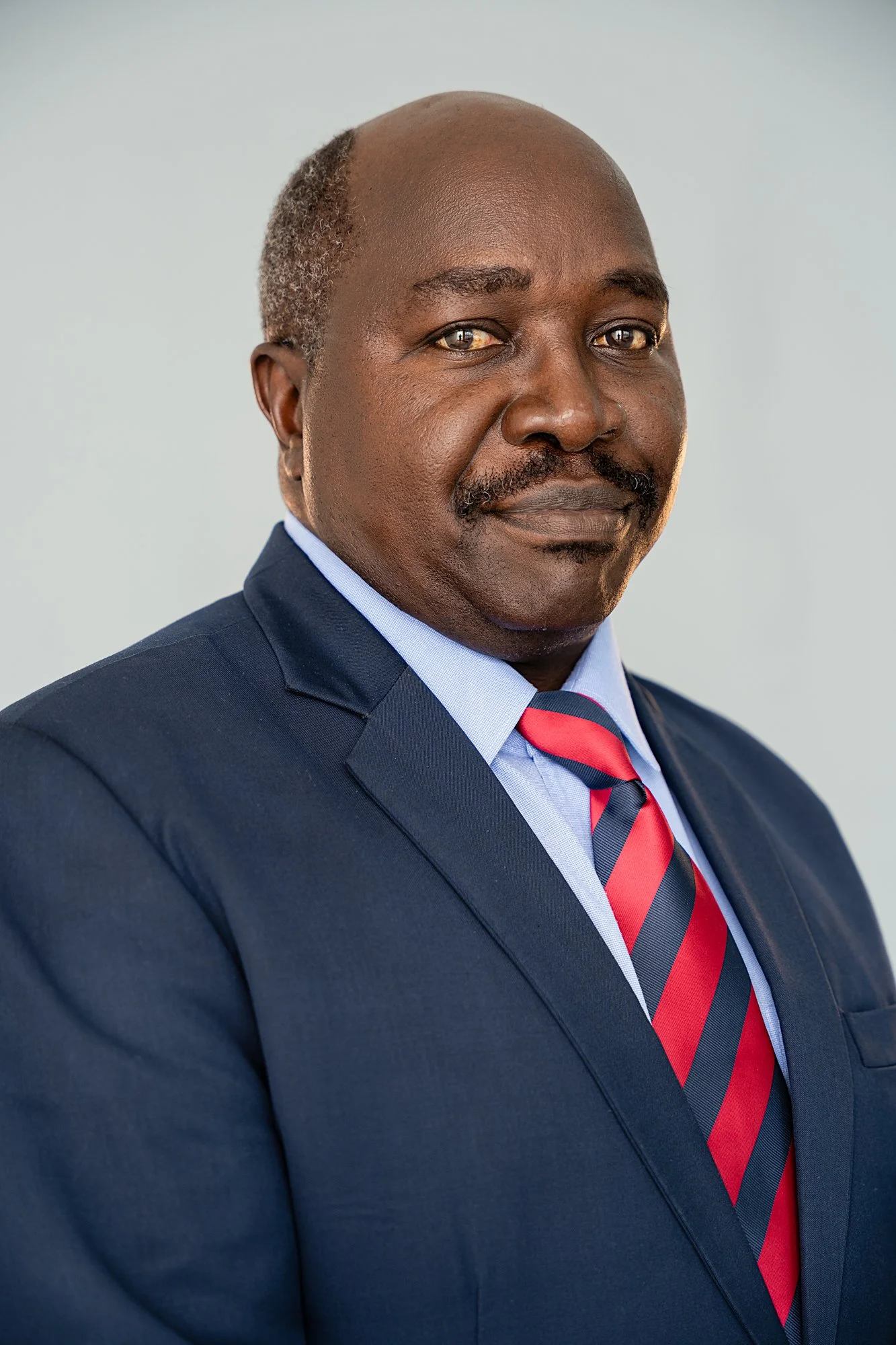 Corporate portrait of a middle-aged African American man in a navy suit, light blue shirt, and red striped tie, looking confidently at the camera against a plain light background.