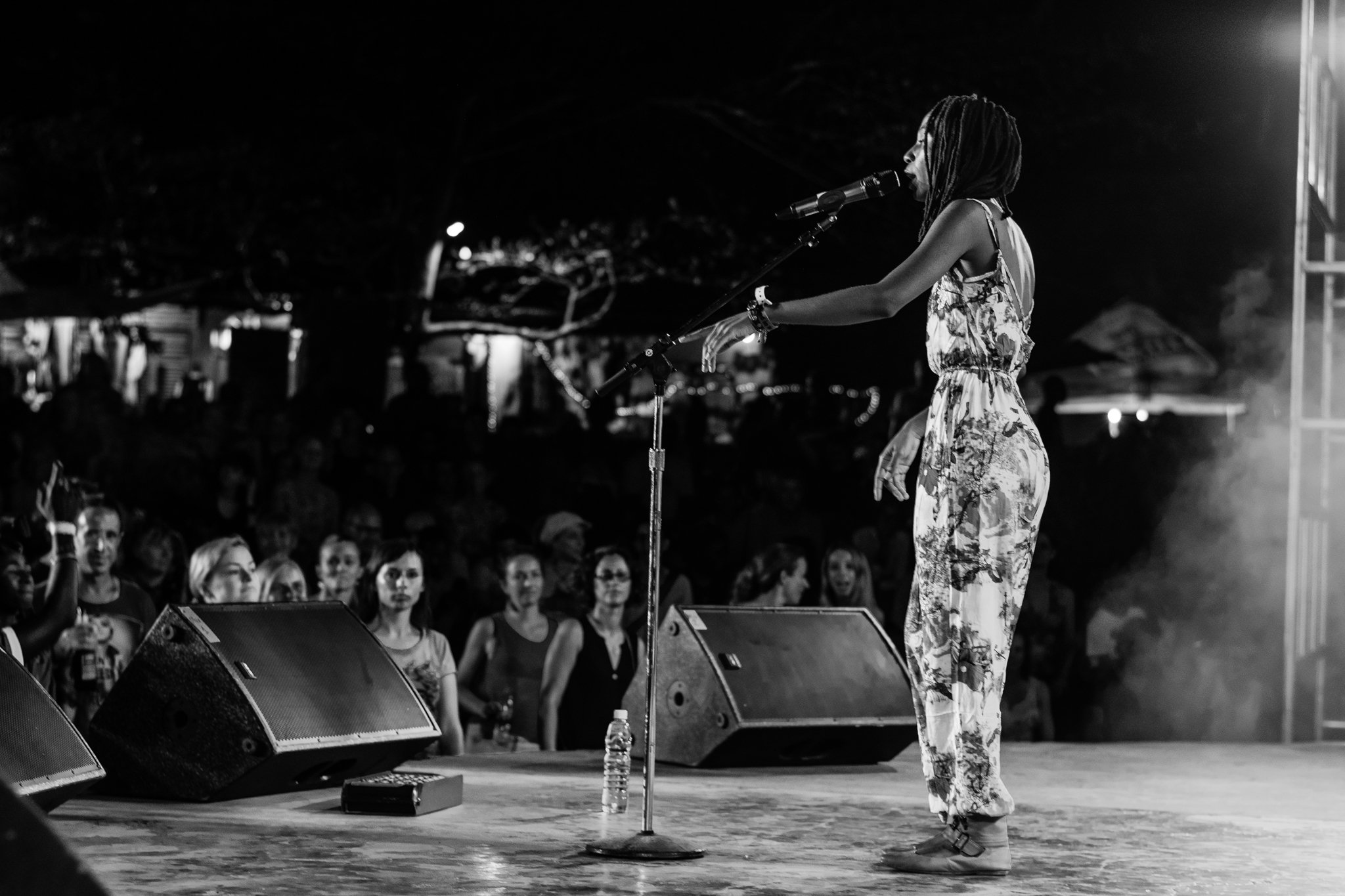 A woman with dreadlocks and a floral dress singing into a microphone on stage at night. Audience members are watching her performance at the Sauti za Busara Music Festival in Zanzibar in 2018.