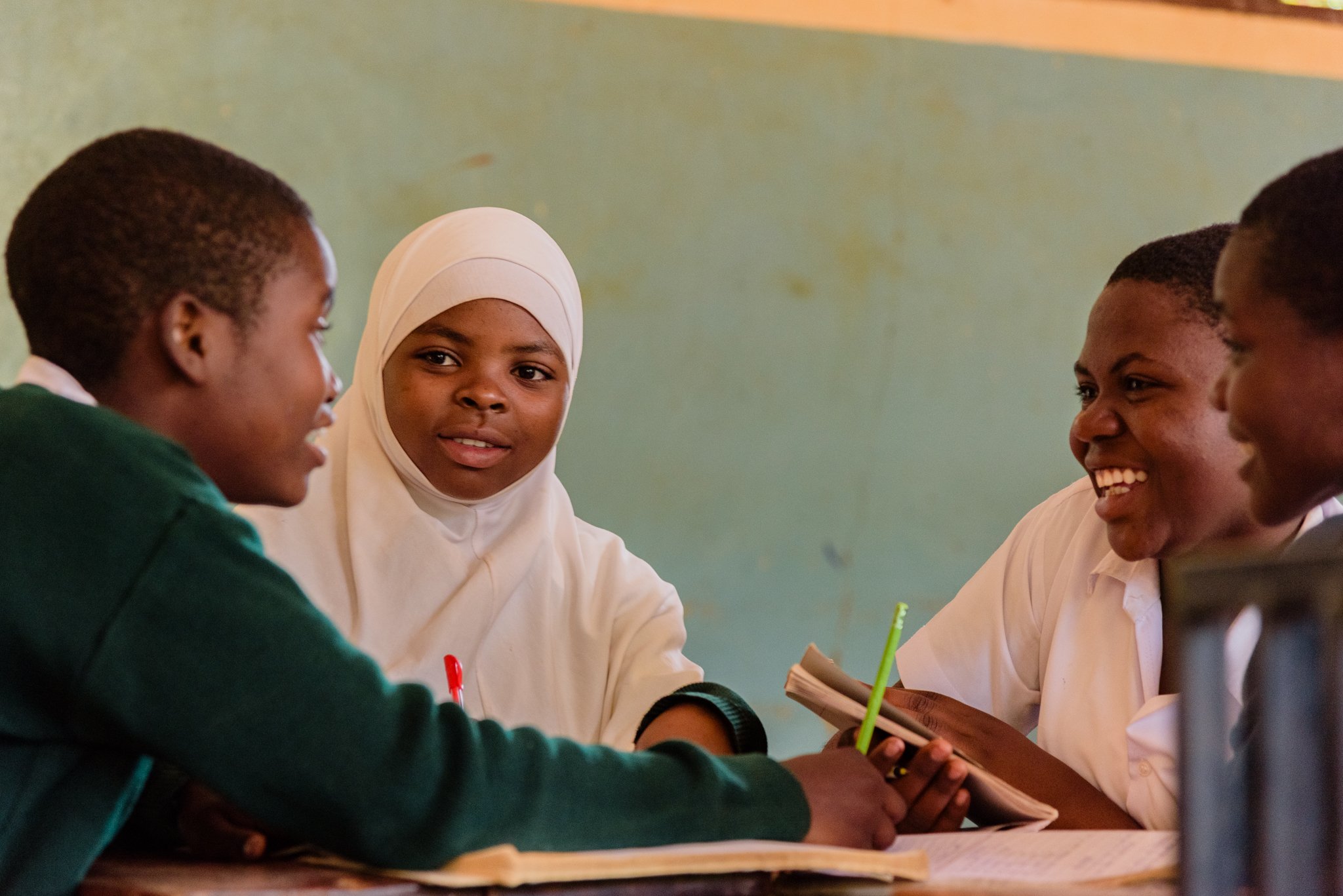 Four female secondary school students sitting at a table engaged in discussion inside a classroom with a green wall in Tanzania, photographed for the Malala Fund girls’ education project.