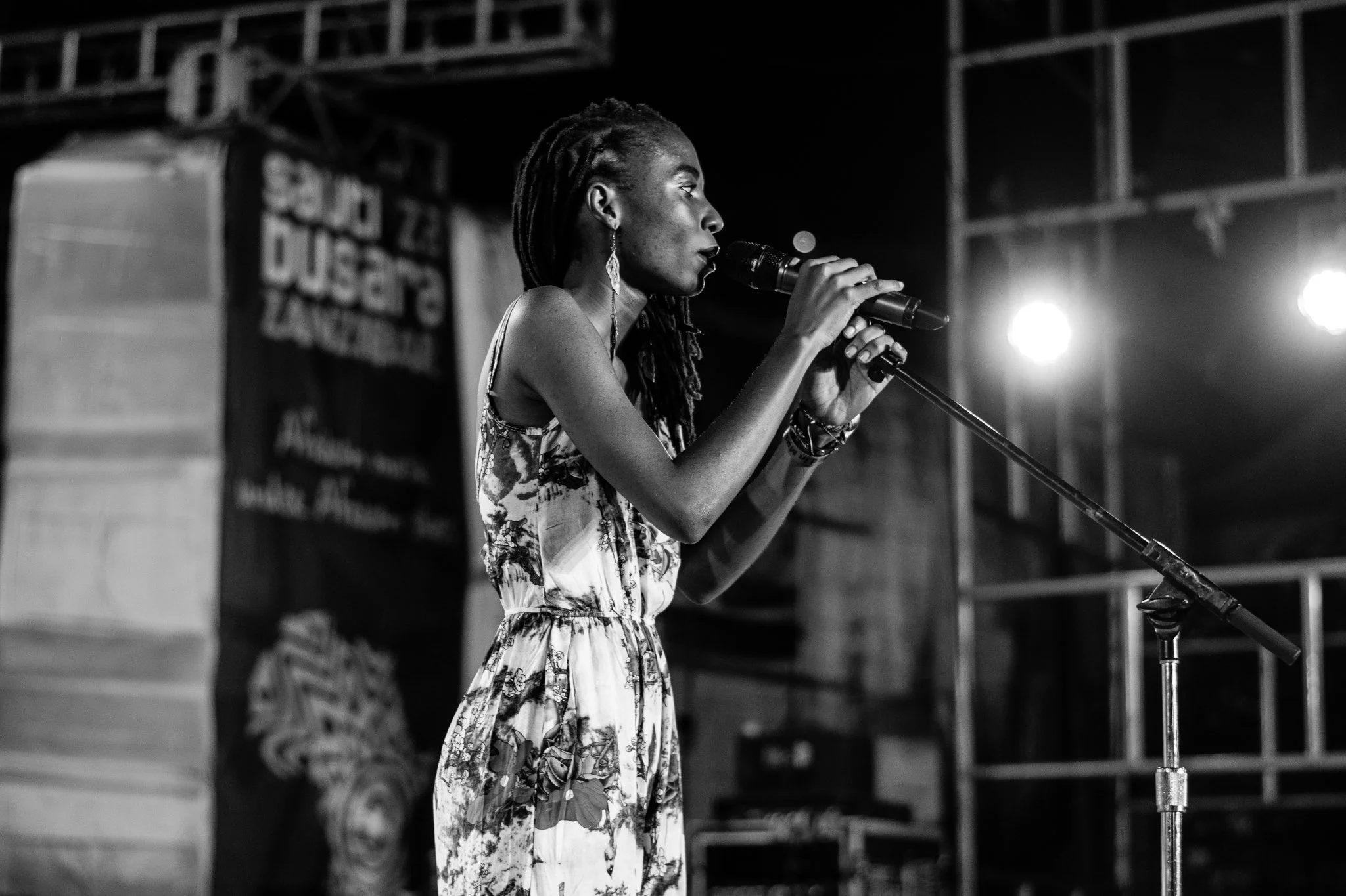 A woman with dreadlocks singing into a microphone on stage, wearing a floral dress, with stage lights in the background, in a black-and-white photograph captured in Zanzibar at the Sauti za Busara Music Festival in 2018.