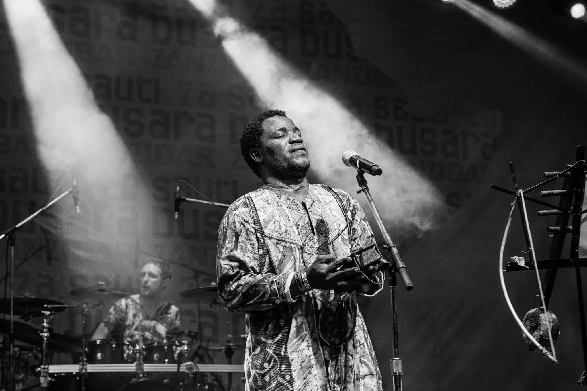 A man is performing on stage, eyes closed, playing a traditional instrument with his hands. A drummer is visible in the background, and stage lights shine down, creating a dramatic atmosphere in Zanzibar at the Sauti za Busara Music Festival