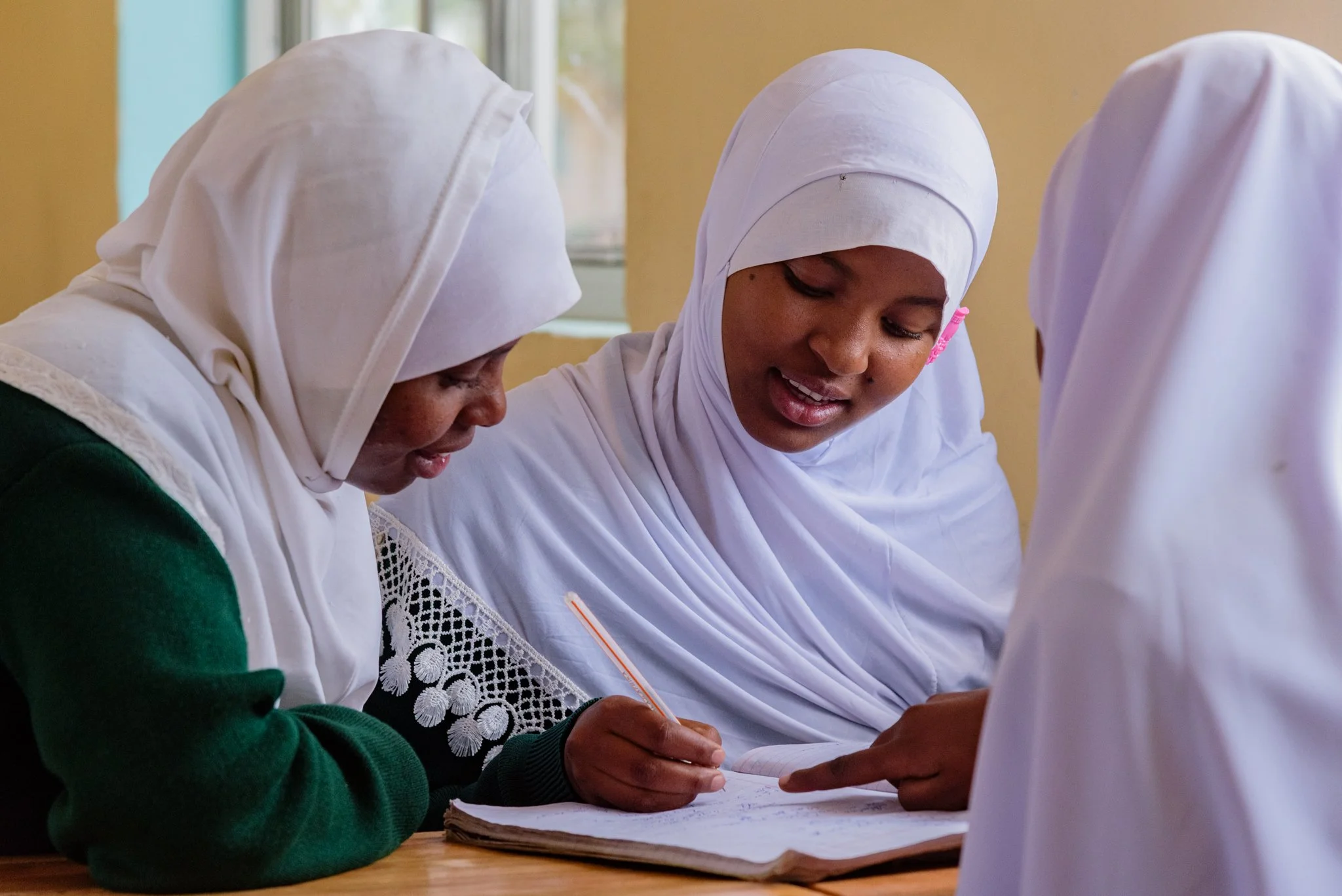 Three Tanzanian schoolgirls wearing white hijabs sitting at desks in a classroom, having a discussion in Dodoma, Tanzania
