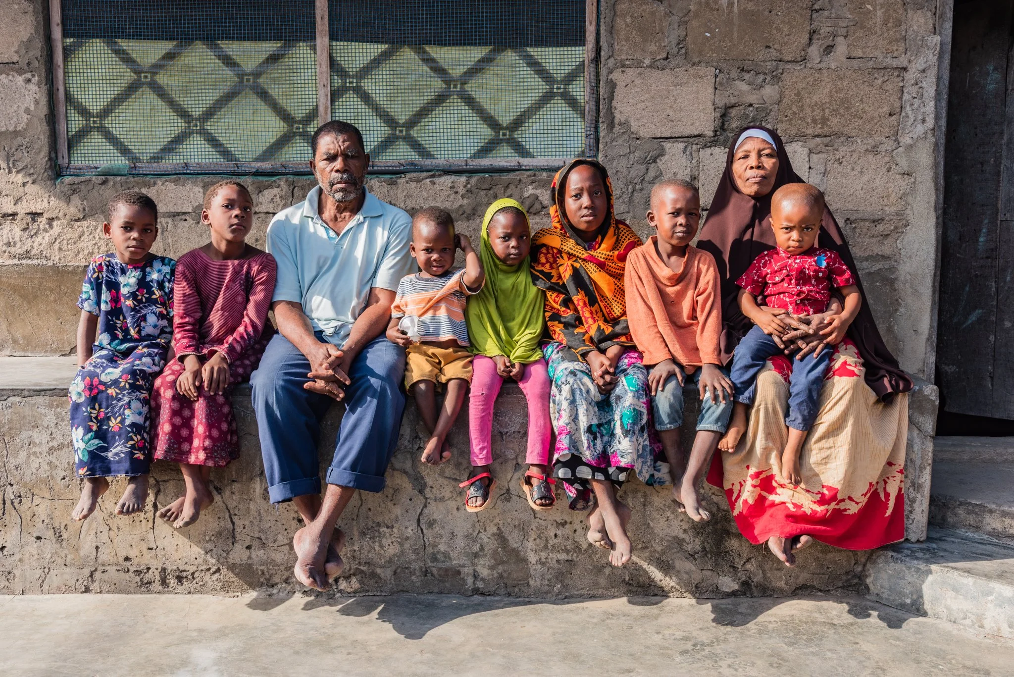 A family of nine people sitting on a concrete ledge outside a textured stone wall, with some children and two adults wearing colorful clothing and barefoot, looking at the camera in Zanzibar, Tanzania.