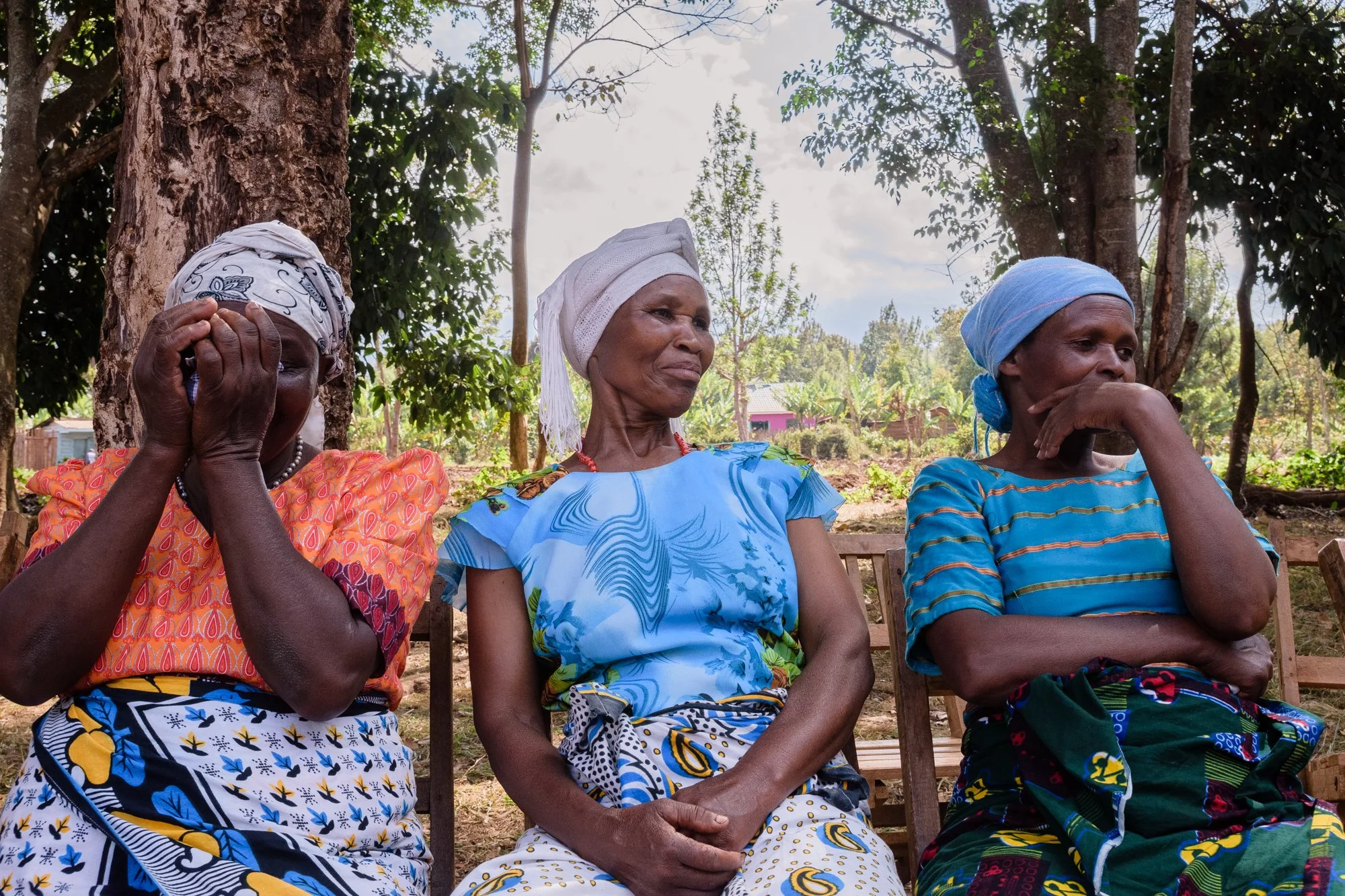 Three elderly women sit on chairs outdoors, wearing colourful traditional clothing and headscarves in Kilimanjaro, Tanzania.