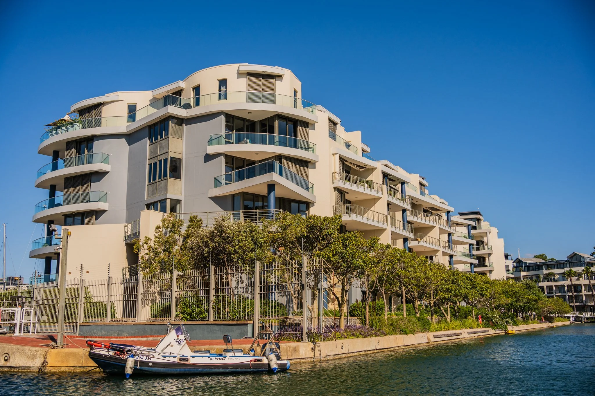 Modern multi-story apartment building by a waterway with a small boat docked at the edge, trees along the fence, and a clear blue sky.