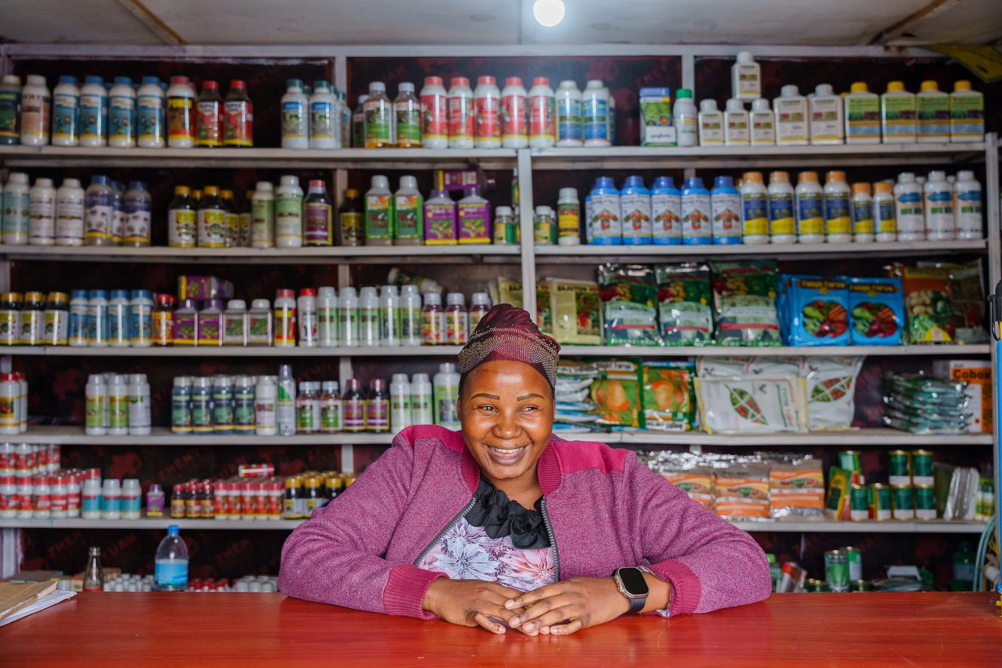 A woman smiling behind the counter of a rural agro-input shop with shelves of fertilizers and seeds in Babati, Manyara, Tanzania.