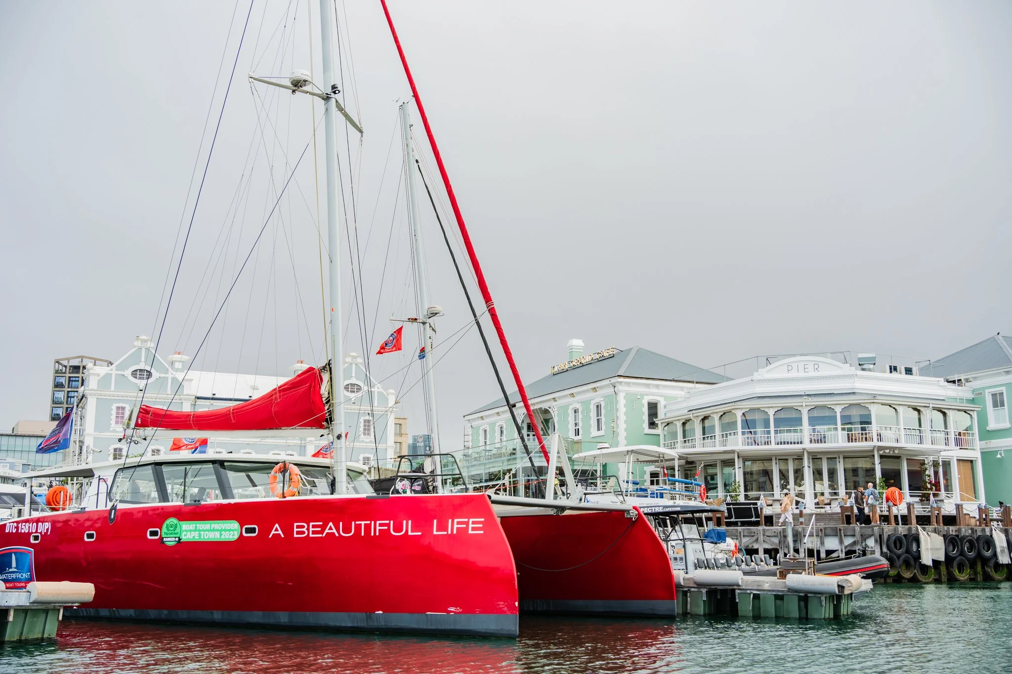 A red sailboat docked at a pier in front of pastel-colored buildings, with a cloudy sky overhead.
