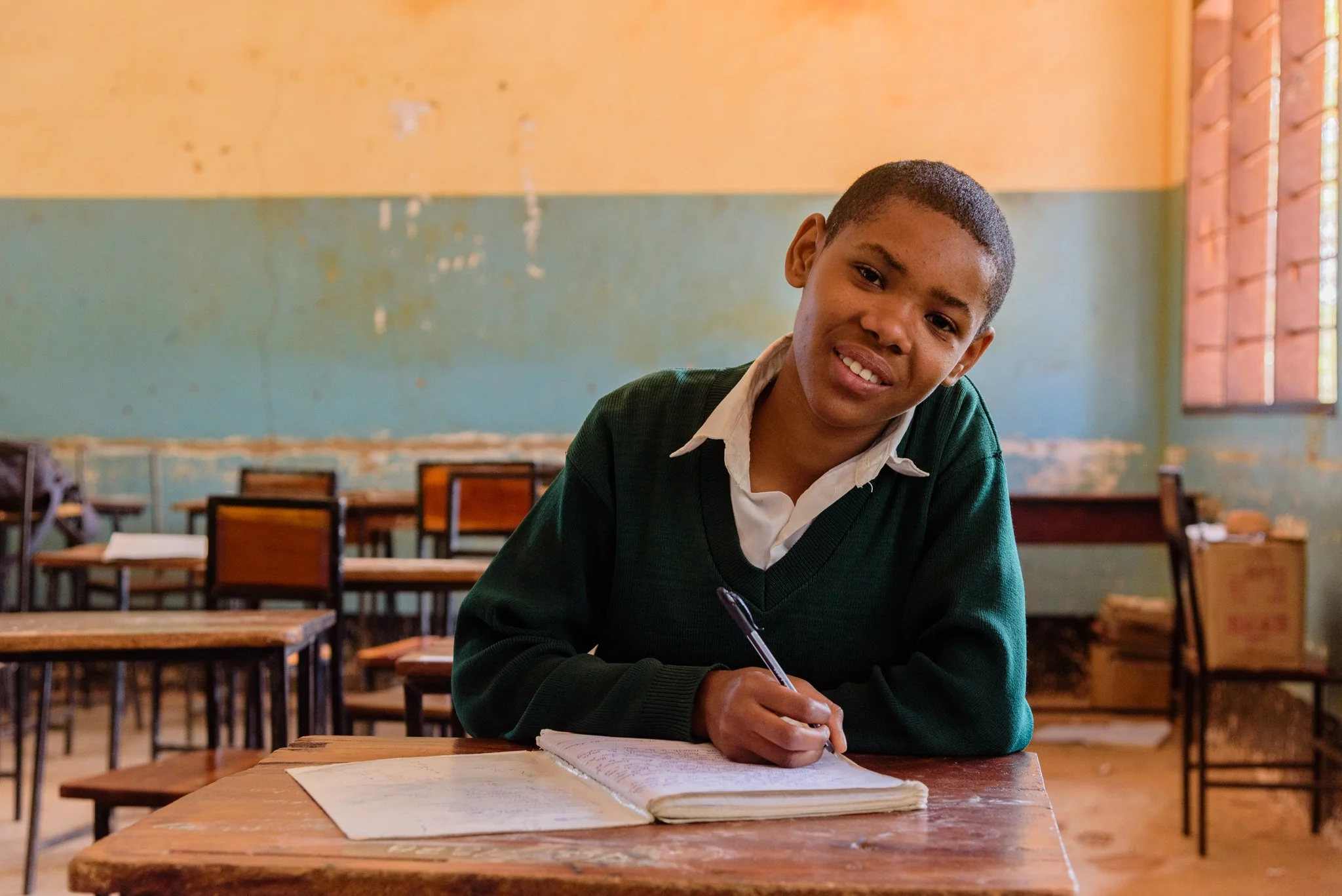 Secondary school student in uniform writing in her notebook at a wooden desk inside a classroom in Dodoma, Tanzania, captured during a Malala Fund assignment documenting girls’ education.