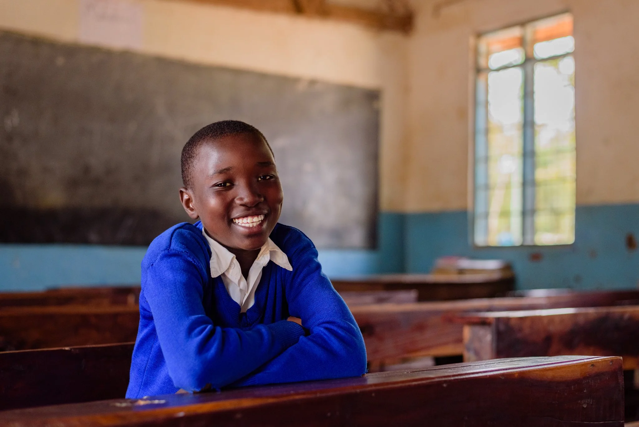 Primary school student smiling at her desk in a classroom with natural light and a blackboard in Dodoma, Tanzania, captured during a Malala Fund assignment focused on girls’ education.