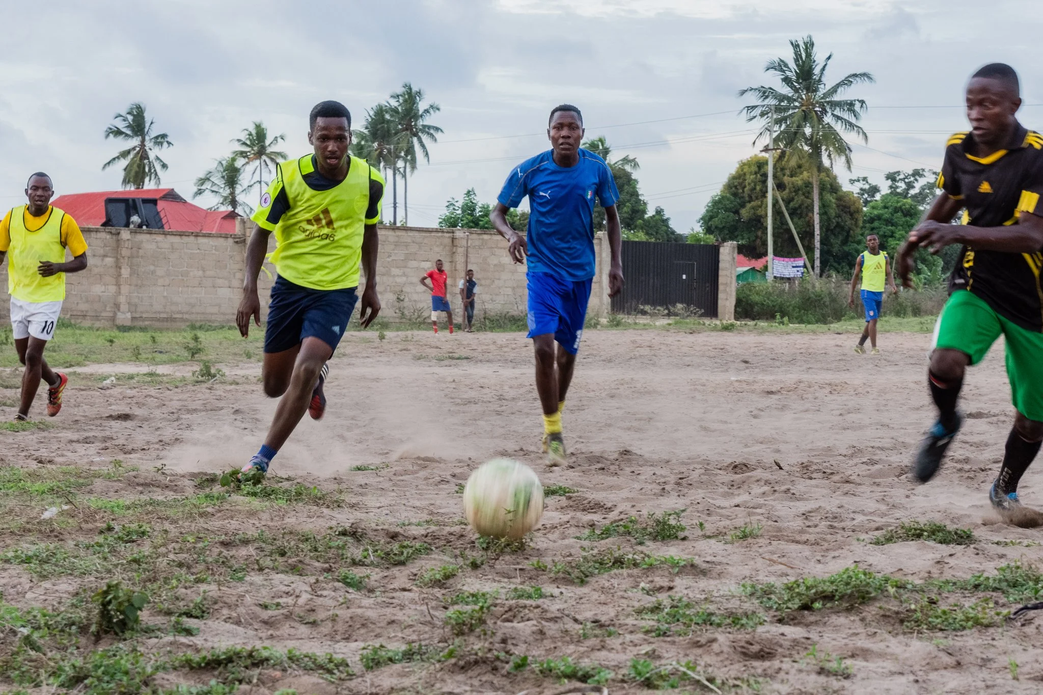 Young men playing football on a sandy field with palm trees and a brick wall in the background in Toangoma, Dar es Salaam.