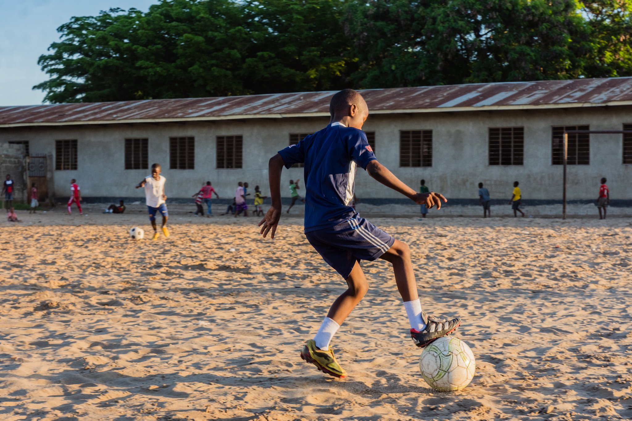 A boy in a blue sports uniform playing football on a sandy field with children in the background and a school building behind them in Mbagala, Dar es Salaam.