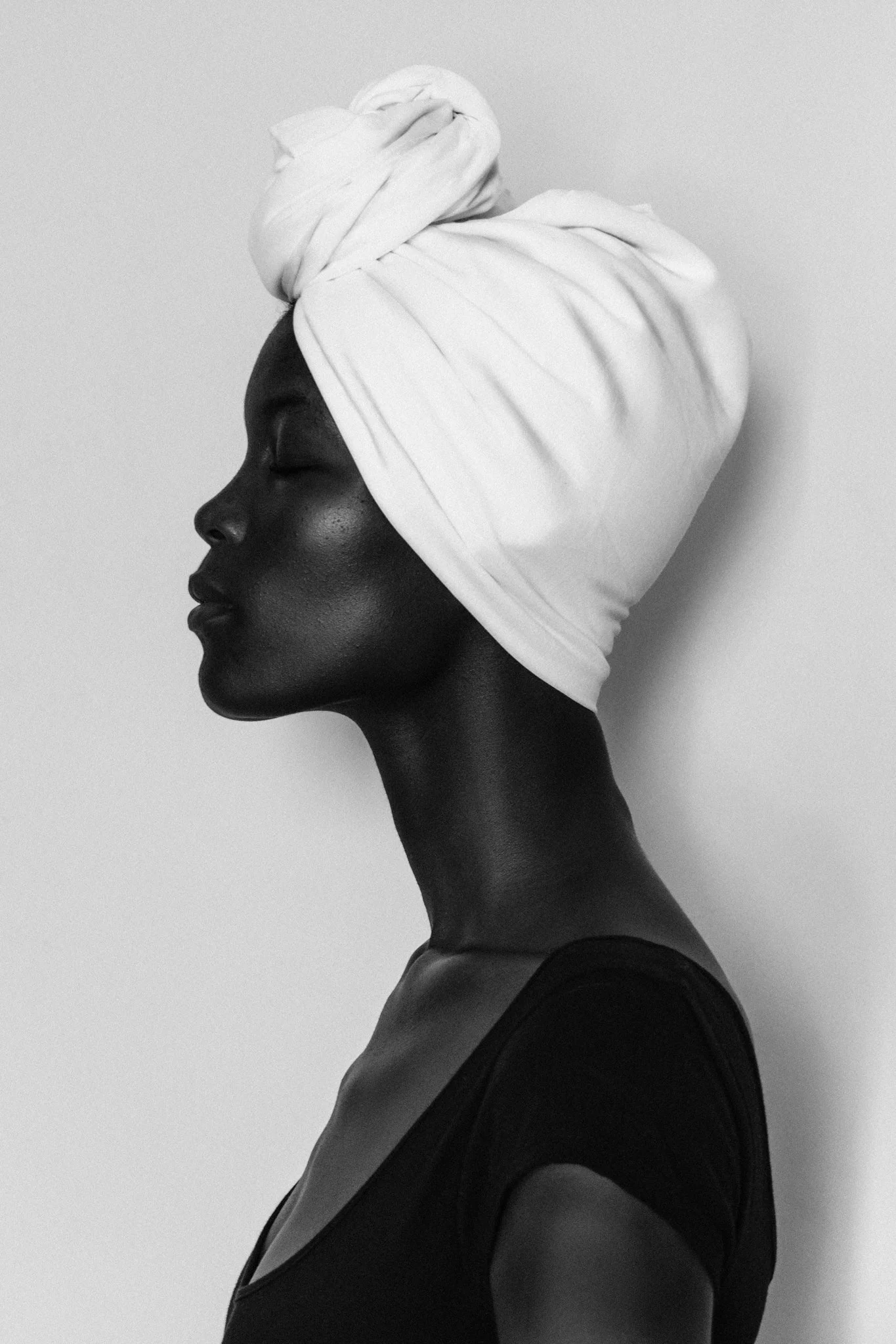 A high-contrast, black and white fine art side-profile portrait of a female model with a white headwrap, facing to the left, with her eyes closed and wearing a black top in Dar es Salaam, Tanzania.