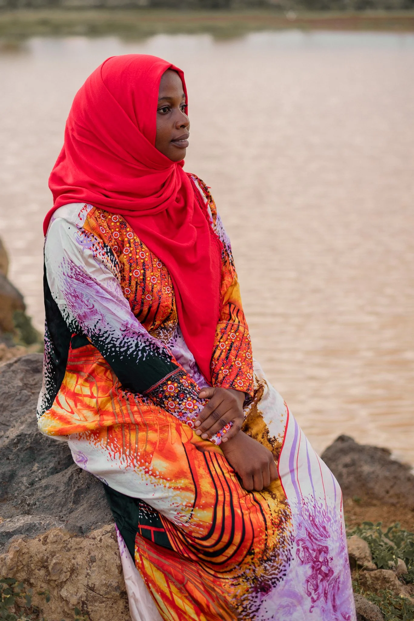 A woman wearing a colorful dress and red headscarf sitting on rocks near a body of water.