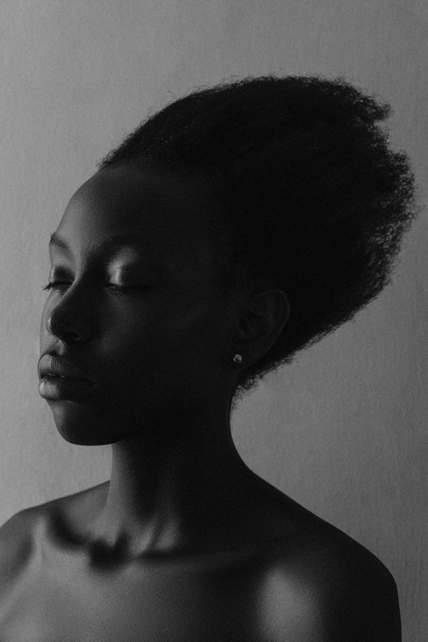A high-contrast, black and white fine art portrait of a black female model with short curly hair, her eyes closed, and no clothing visible except earrings.