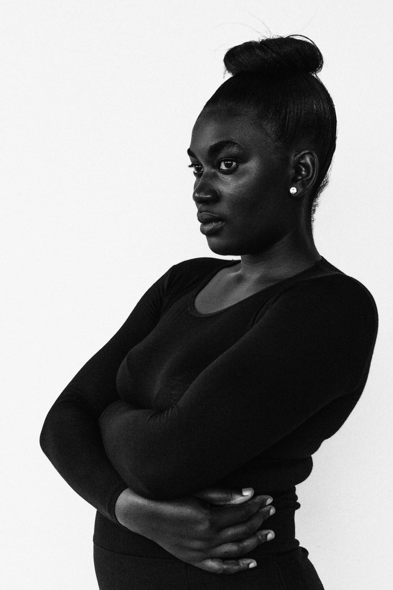 A high-contrast, black-and-white fine art photo of a woman with her arms crossed, wearing a long-sleeve top and earrings, looking to the left, against a plain light background.