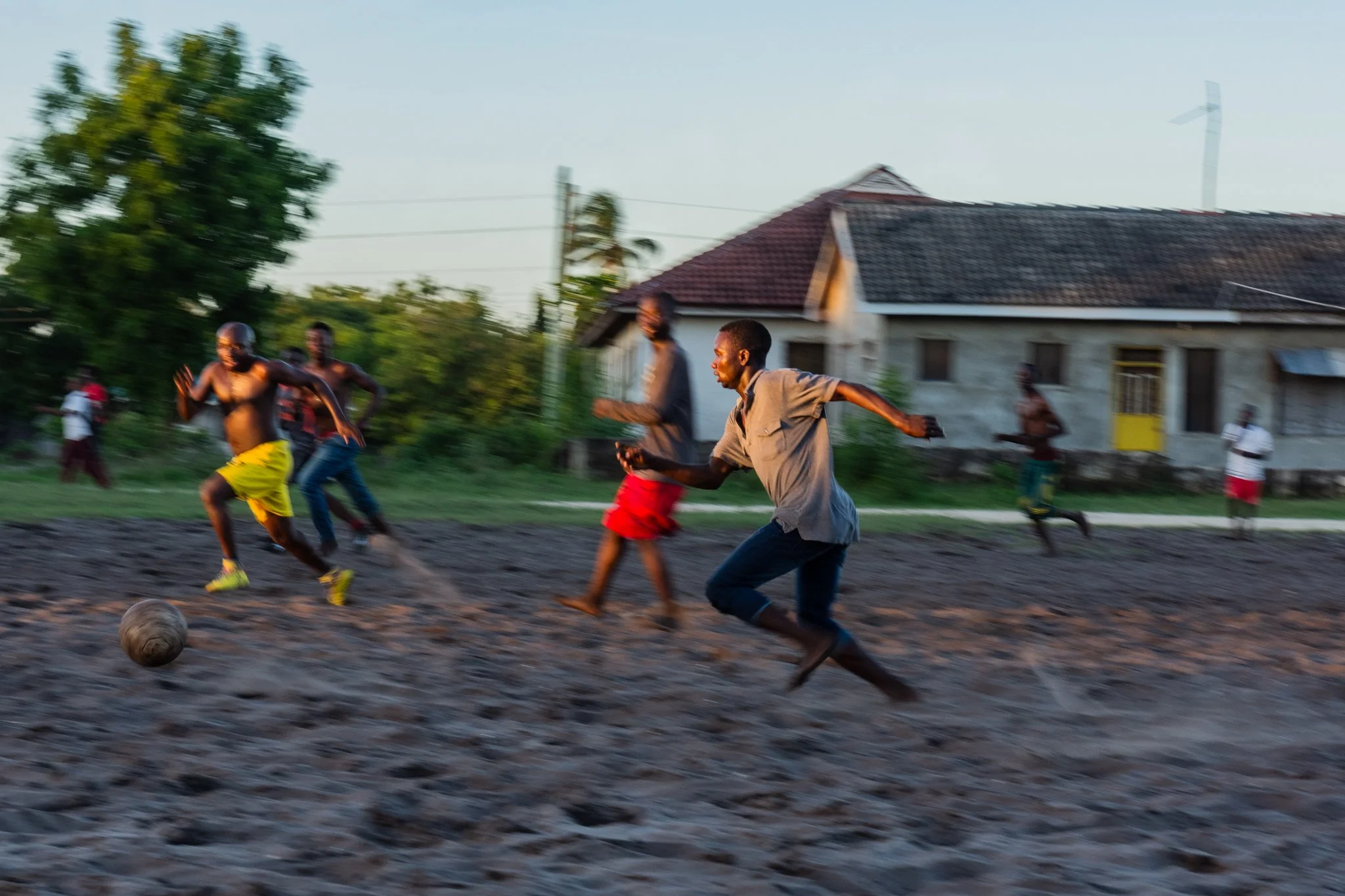 Men playing football on a sandy field with houses and trees in the background during late afternoon in Kigamboni, Dar es Salaam.