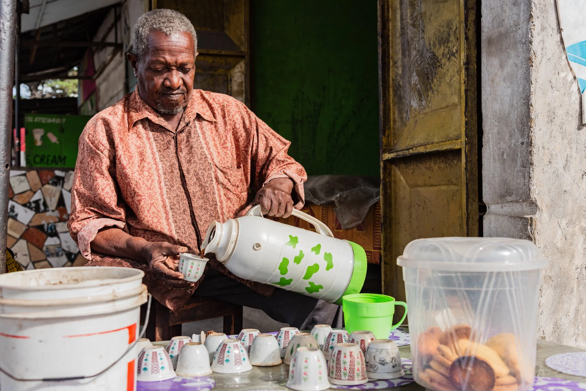 An elderly man pouring coffee from a thermos into a small cup at an outdoor stall, with cups, a plastic container filled with baked goods, and other utensils on the table in Zanzibar, Tanzania.