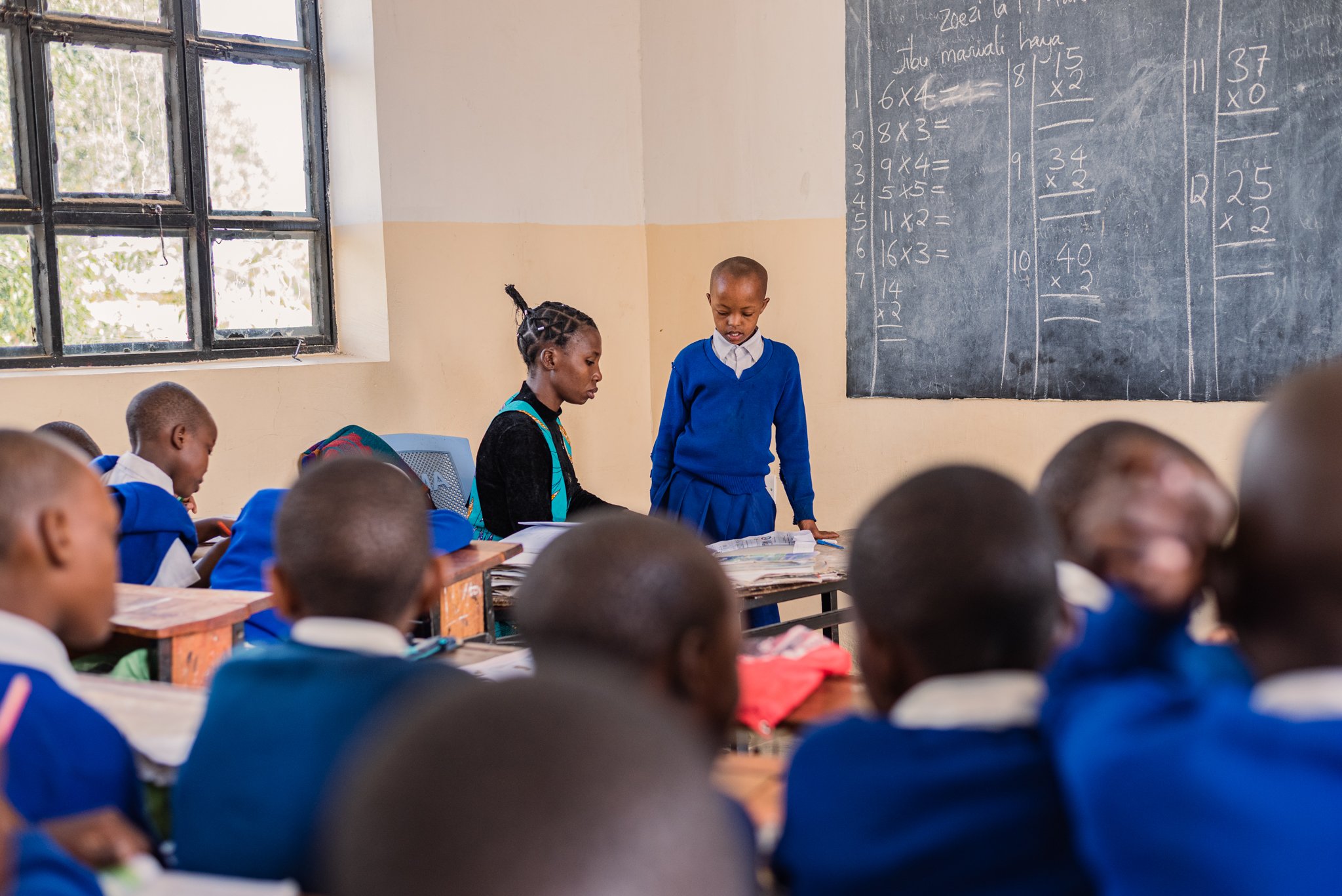 A classroom with students in blue uniforms sitting at desks, a teacher assisting a student at the front, and a blackboard with math problems on the wall in Dodoma, Tanzania.