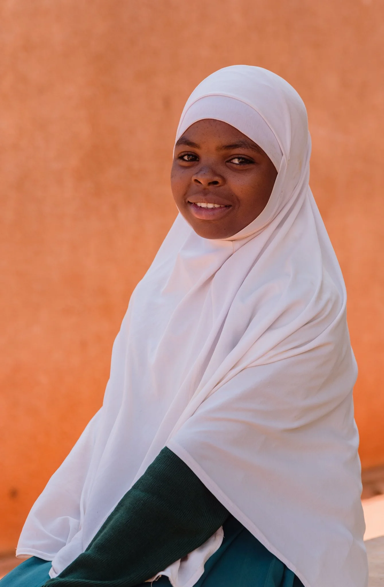 Secondary school student in a white hijab and green uniform standing against an orange wall in Tanzania, captured during a Malala Fund assignment focused on girls’ education.