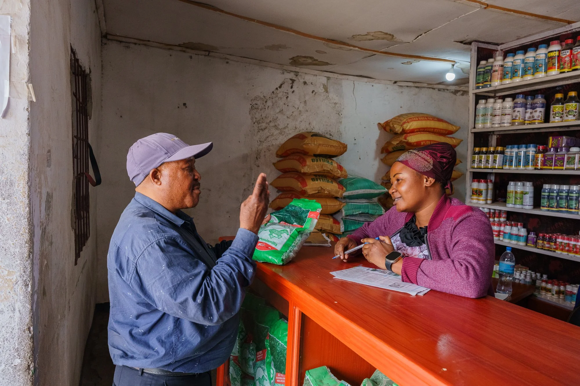 A man and a woman speaking at a shop counter with shelves of agricultural supplies behind them during a Farm Africa and Farm to Market Alliance project in Babati, Manyara, Tanzania.