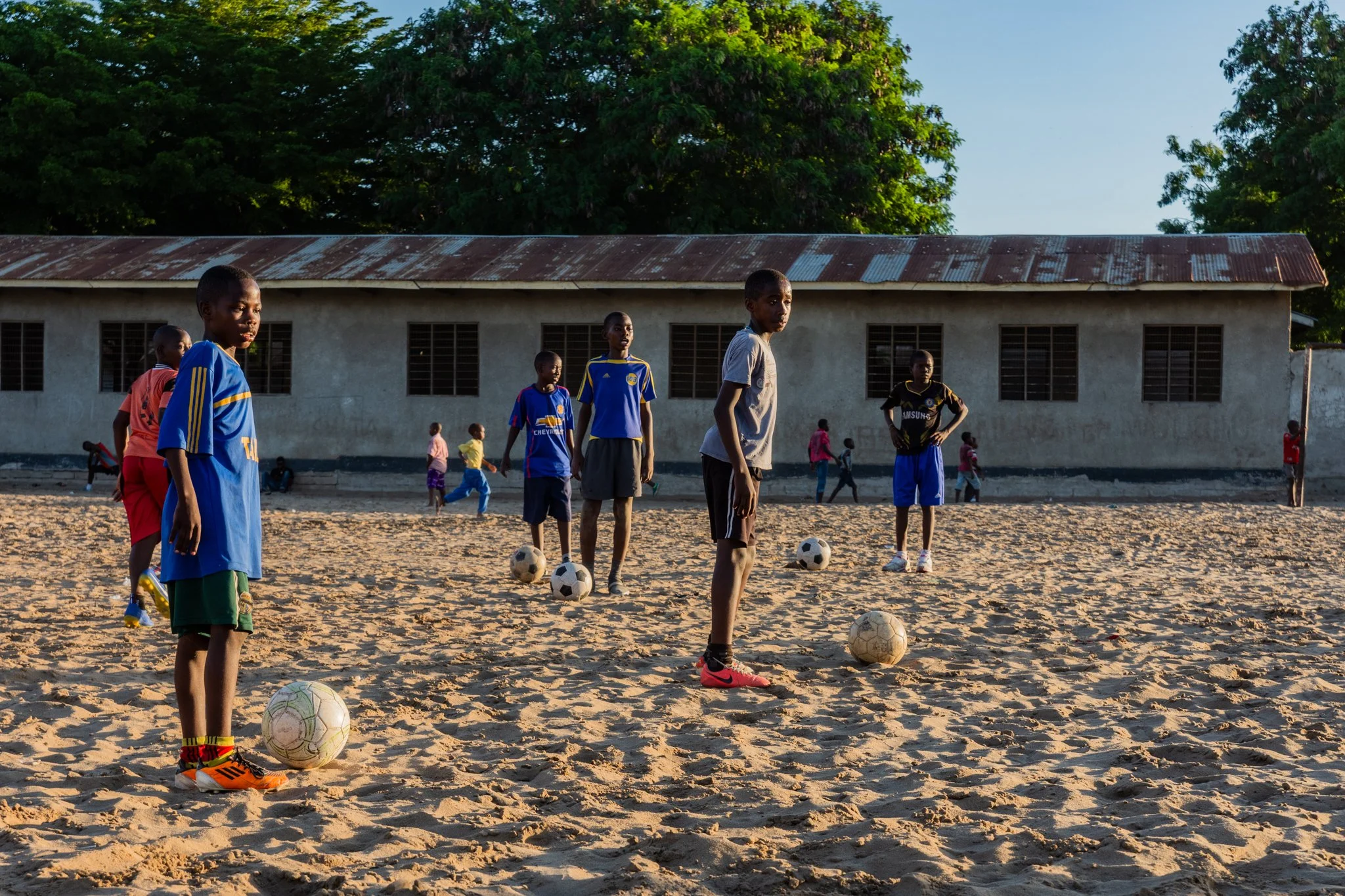 Children practising football on a sandy field with several balls and a school building with trees in the background in Mbagala, Dar es Salaam.