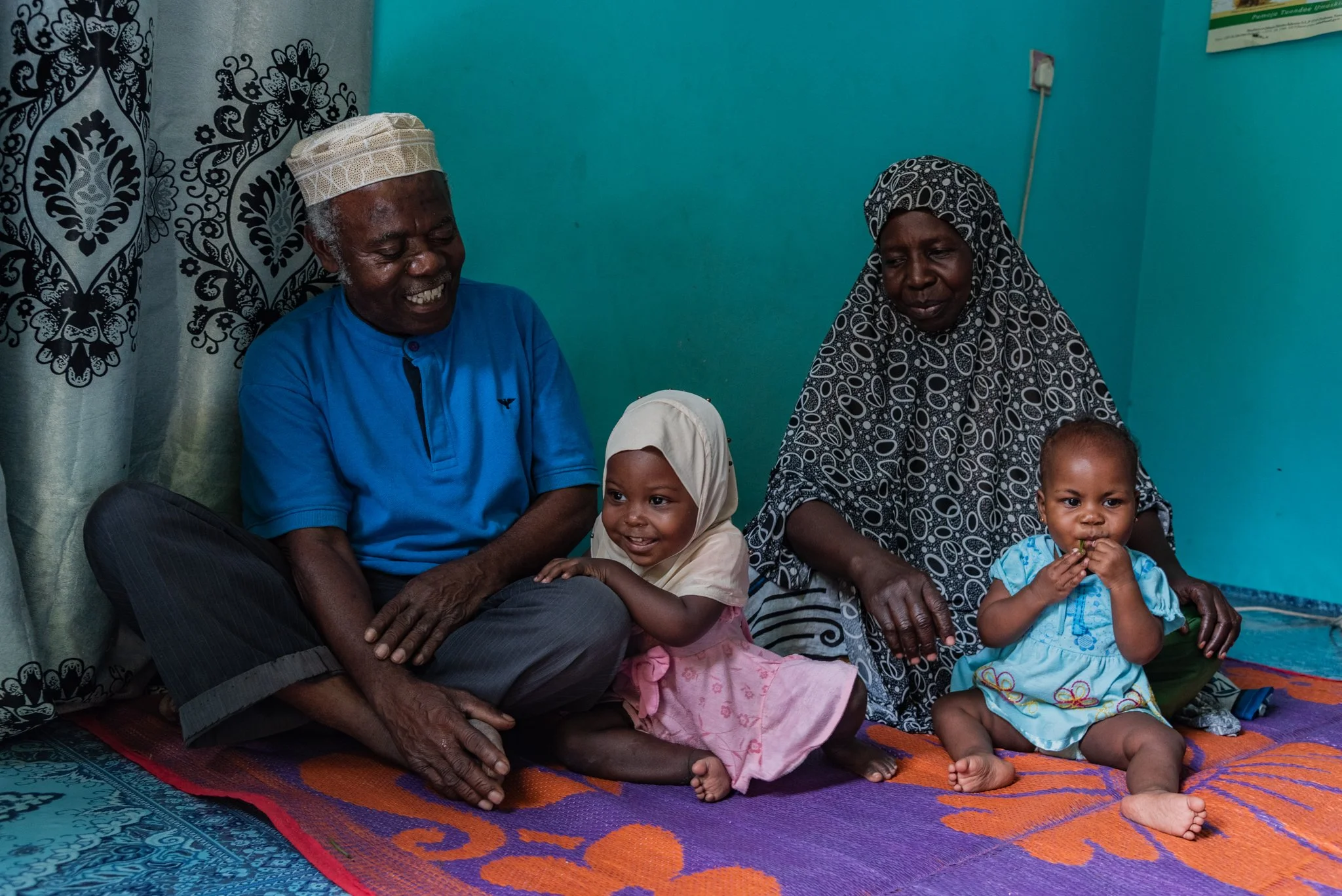 An elderly man and woman sit on the floor with two young children, smiling, in a room with bright blue walls and colourful rugs in Zanzibar, Tanzania.