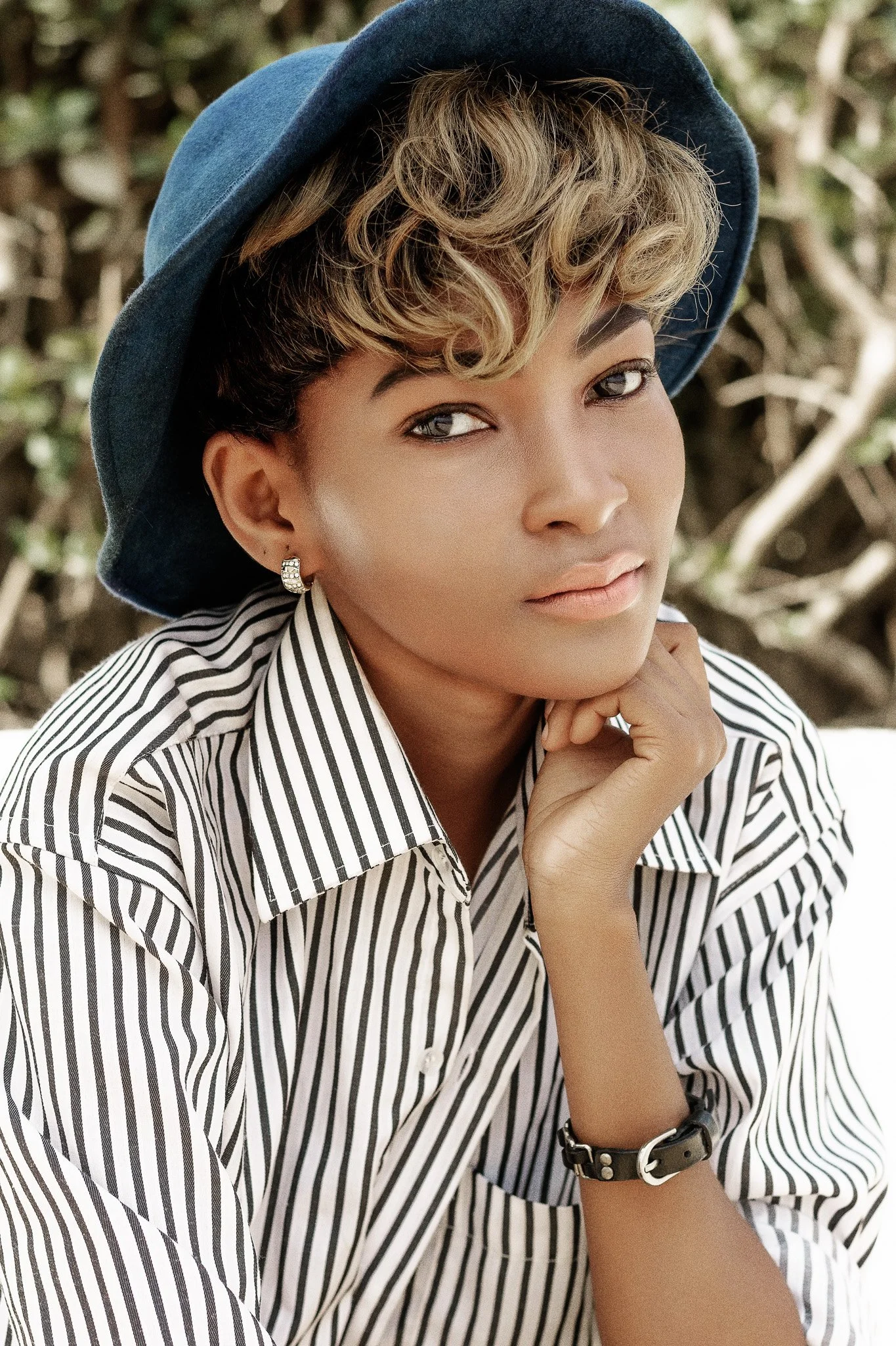 Fashion editorial portrait of a young woman with short curly hair wearing a blue hat and striped shirt, resting her chin on her hand outdoors in Dar es Salaam, Tanzania.