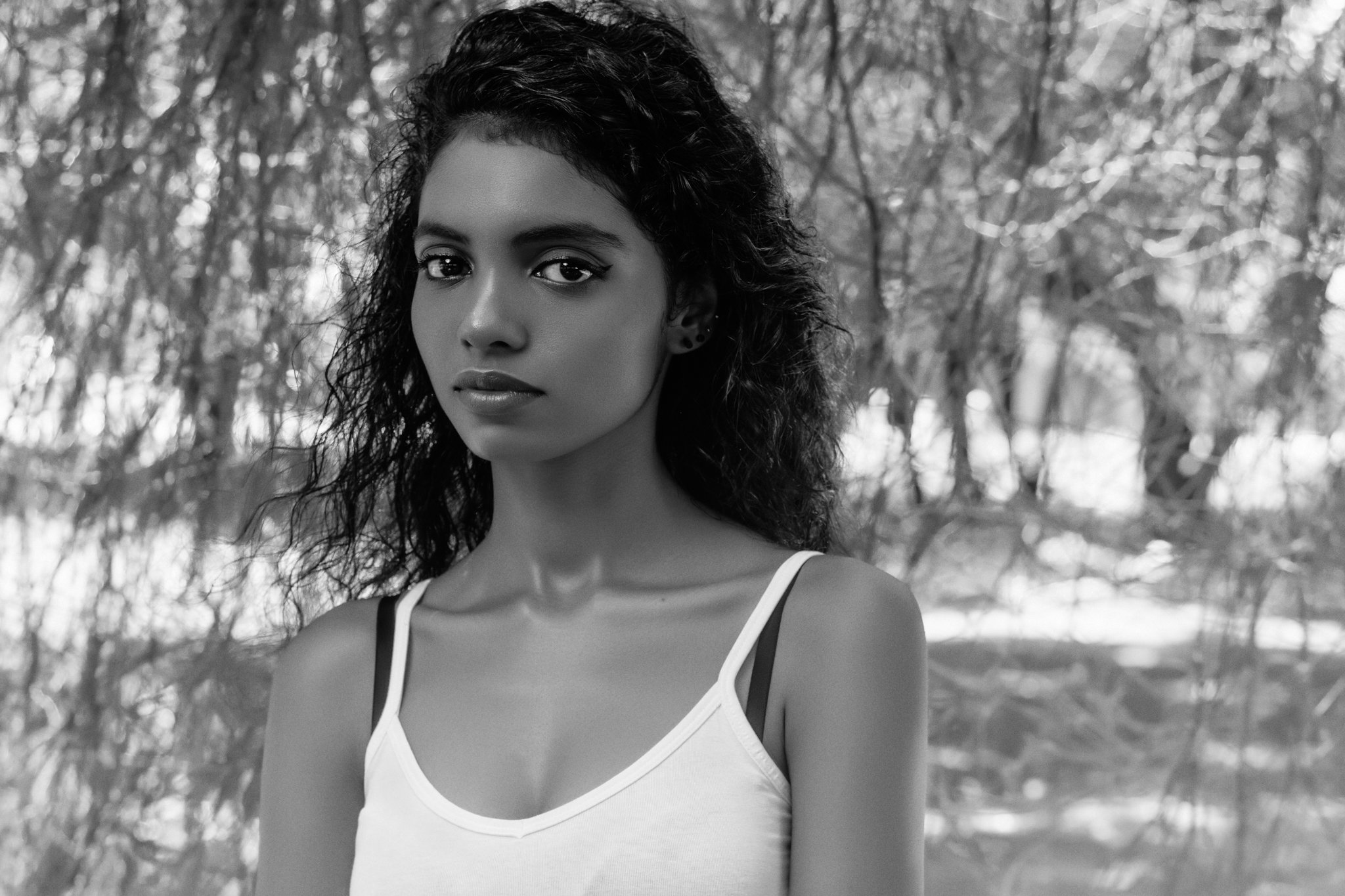 A young woman with curly hair and dark eyes looking directly at the camera, standing outdoors with a background of blurred trees and foliage.
