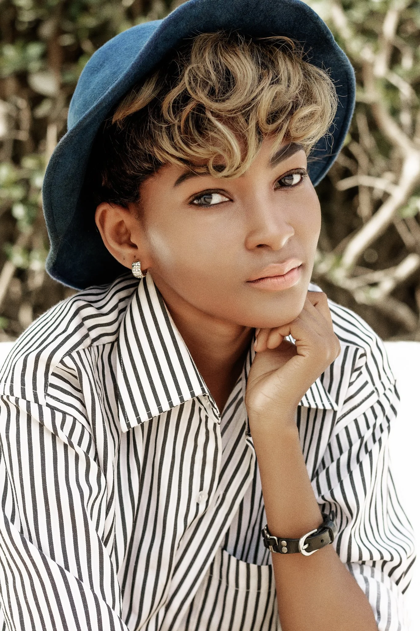 A woman wearing a striped shirt, a black watch, and earrings, posing outdoors with a large blue hat and short, curly hair with highlights.