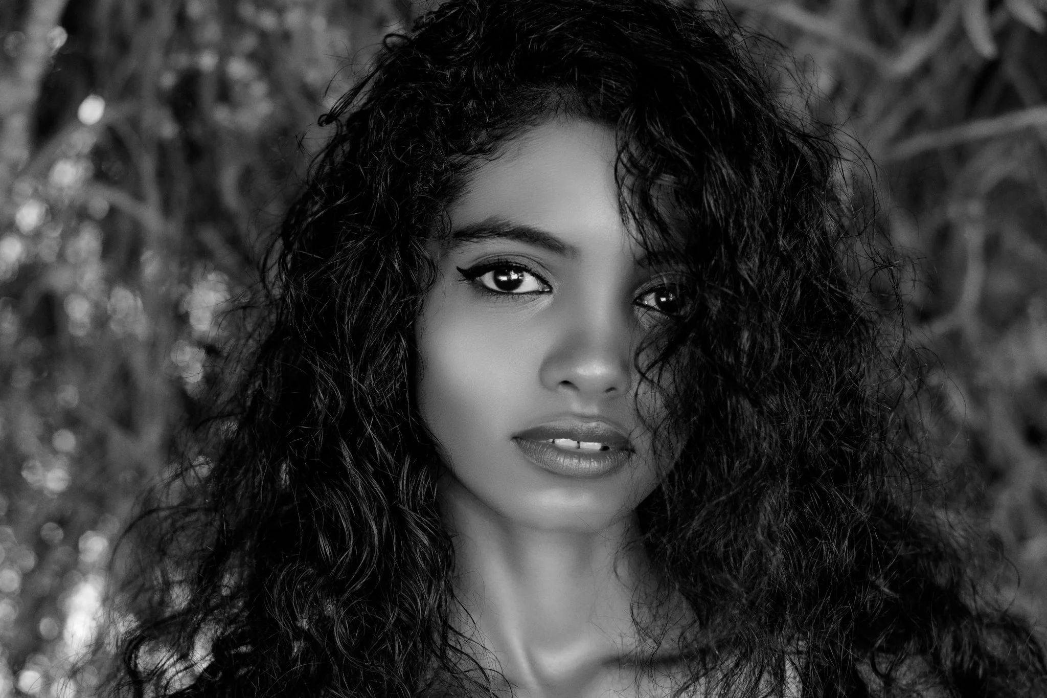 A black and white portrait of a woman with curly hair and intense eyes, looking directly at the camera.