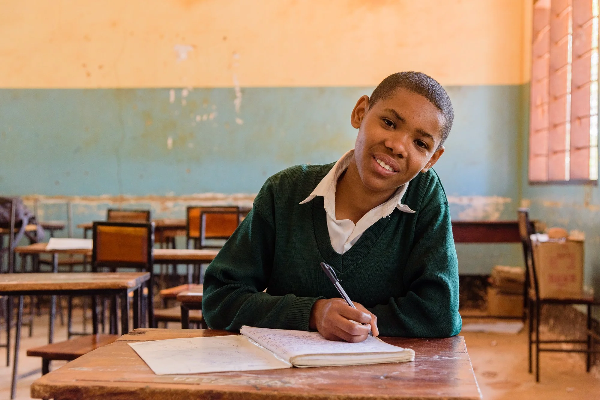 Young girl in a school uniform sits at a wooden desk with an open notebook and pen inside a classroom in Dodoma, Tanzania.