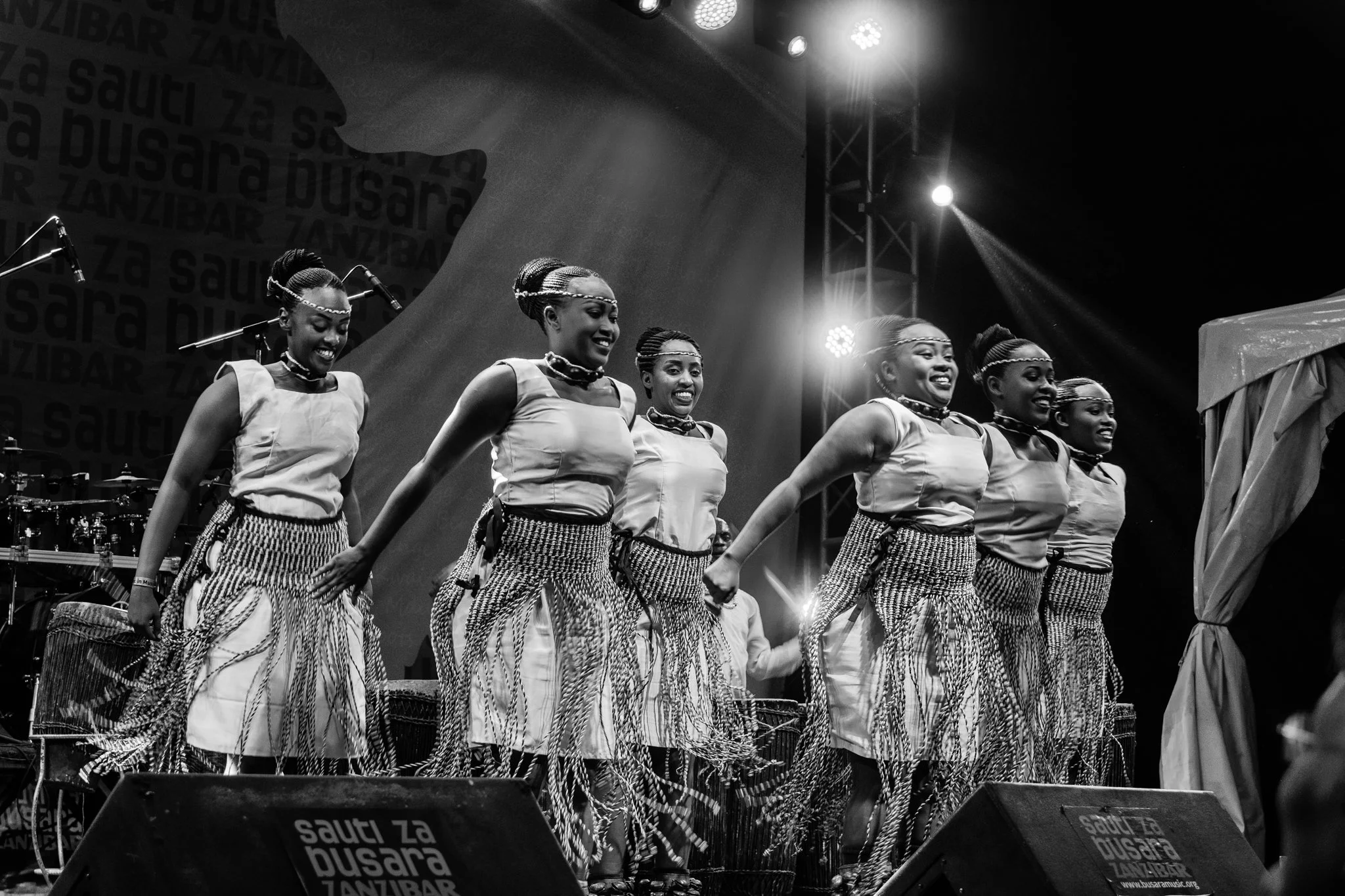 Six women performing on stage with traditional attire, smiling, in front of a backdrop with the text "SAUTI ZA BUSARA", in Zanzibar at the Sauti za Busara Music Festival in 2018.